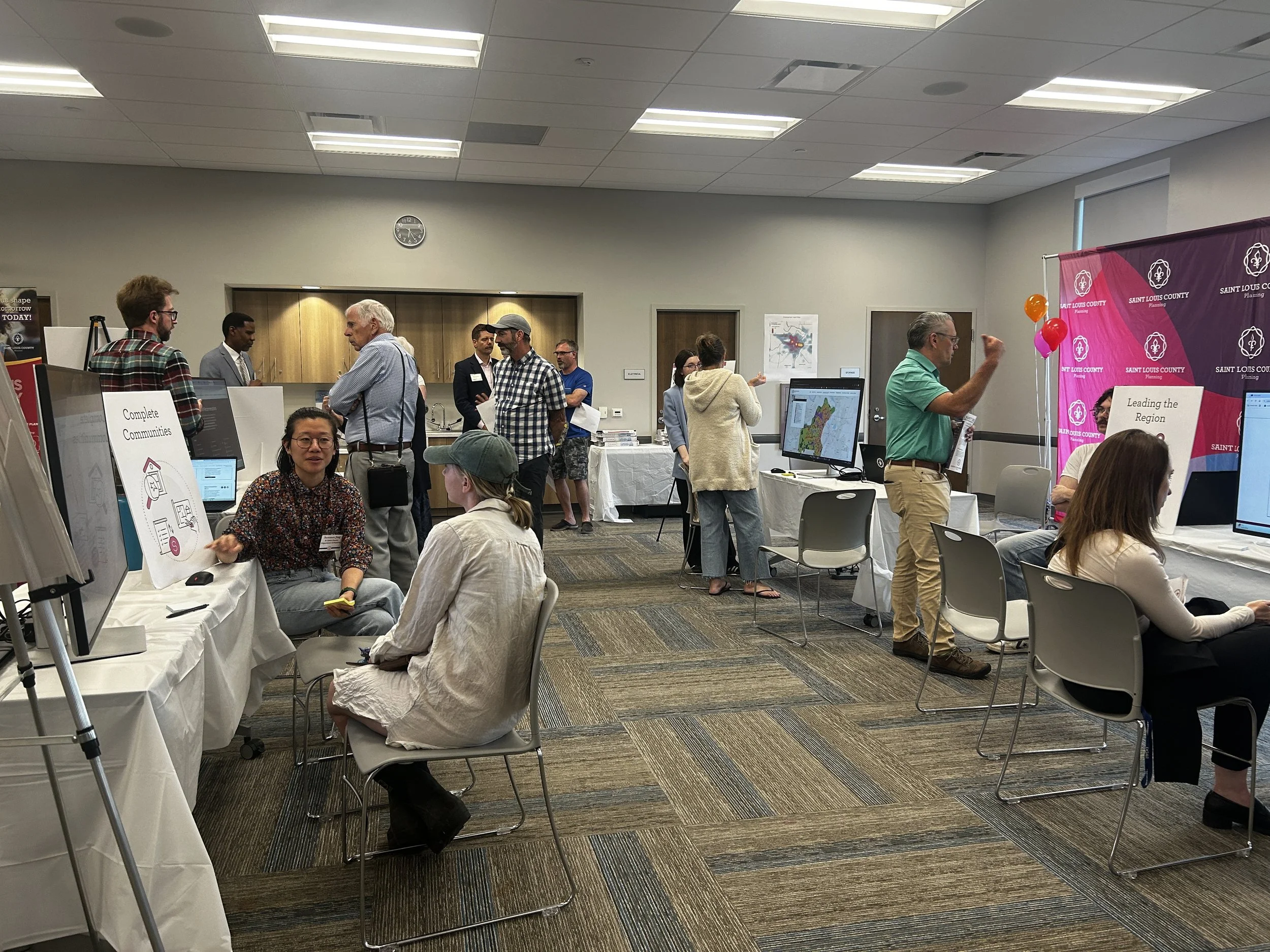 People attending a community event or conference in a room with tables, computers, and informational booths, some engaged in conversations and presentations.