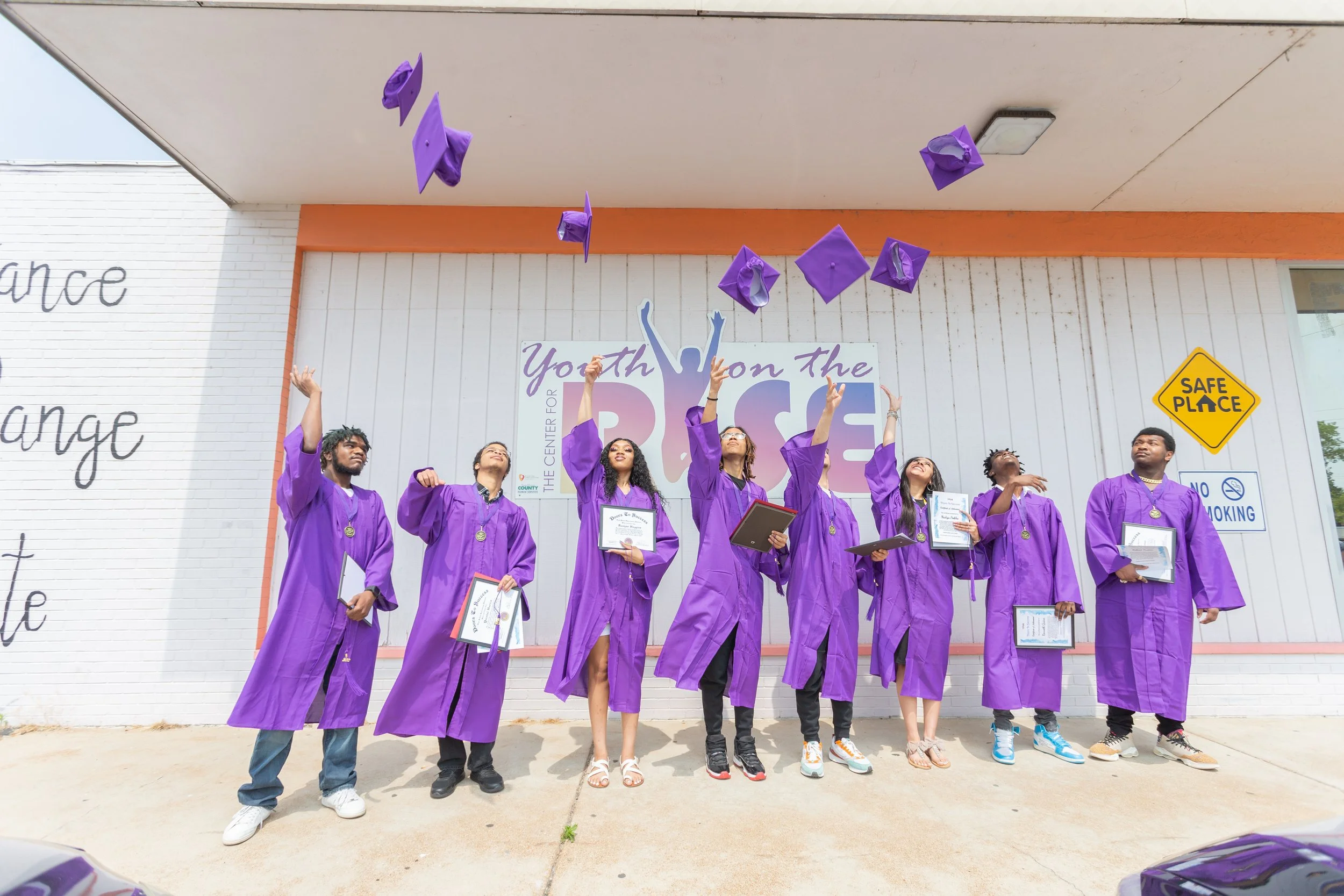 A group of graduates from the Youth on the Rise program stand in purple graduation robes and throw their caps into the air.