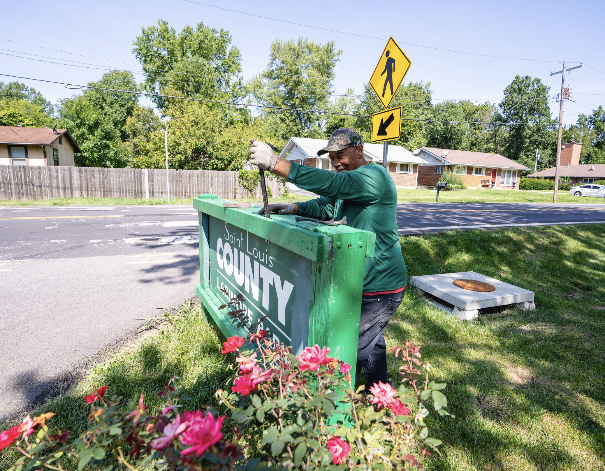 A man working on a street sign that reads 'Saint Louis County' in a suburban neighborhood with houses, trees, and a pedestrian crossing sign in the background.