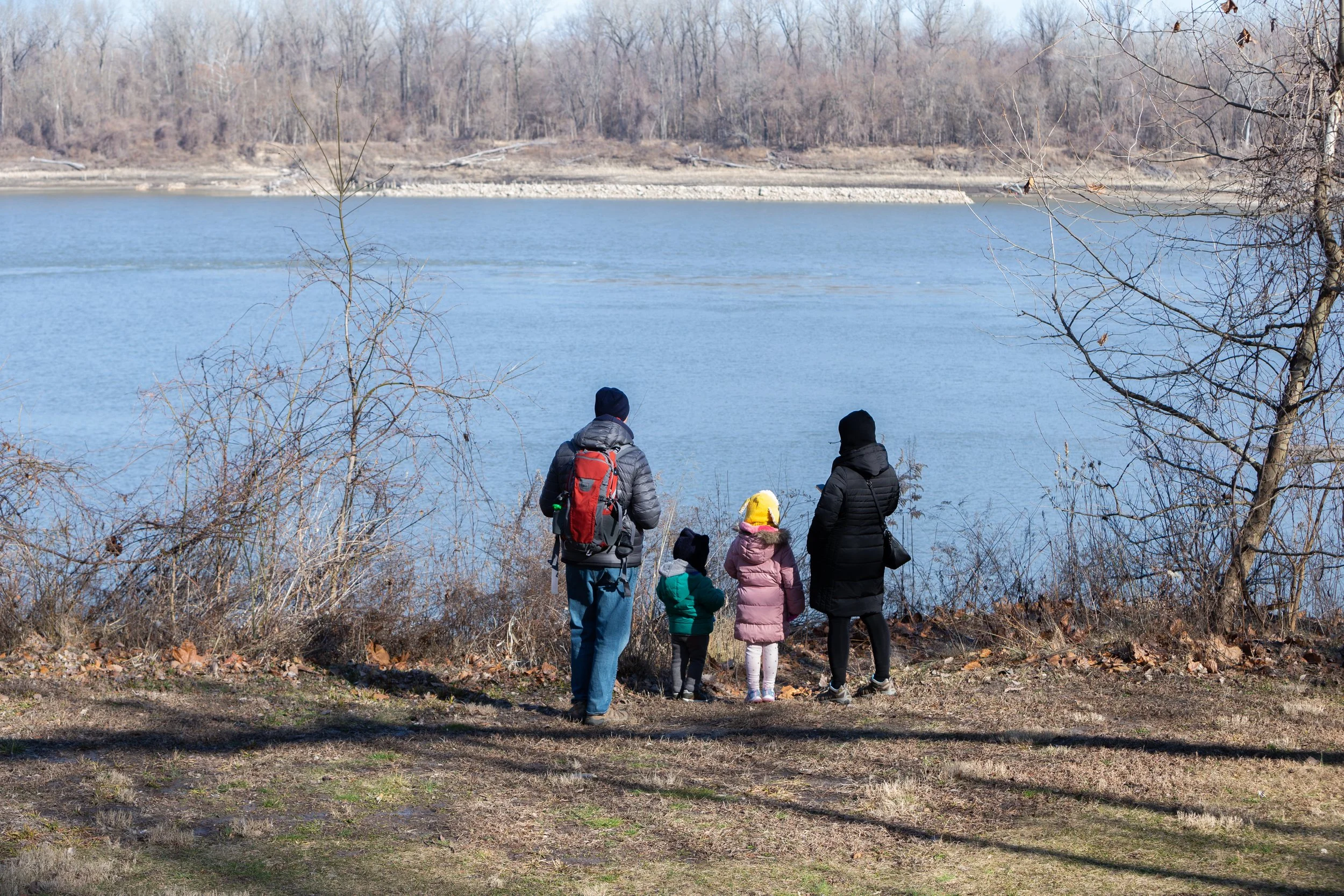 A family stands at the edge of a river in Bellefontaine Park. They are looking across the river with their backs to the camera.