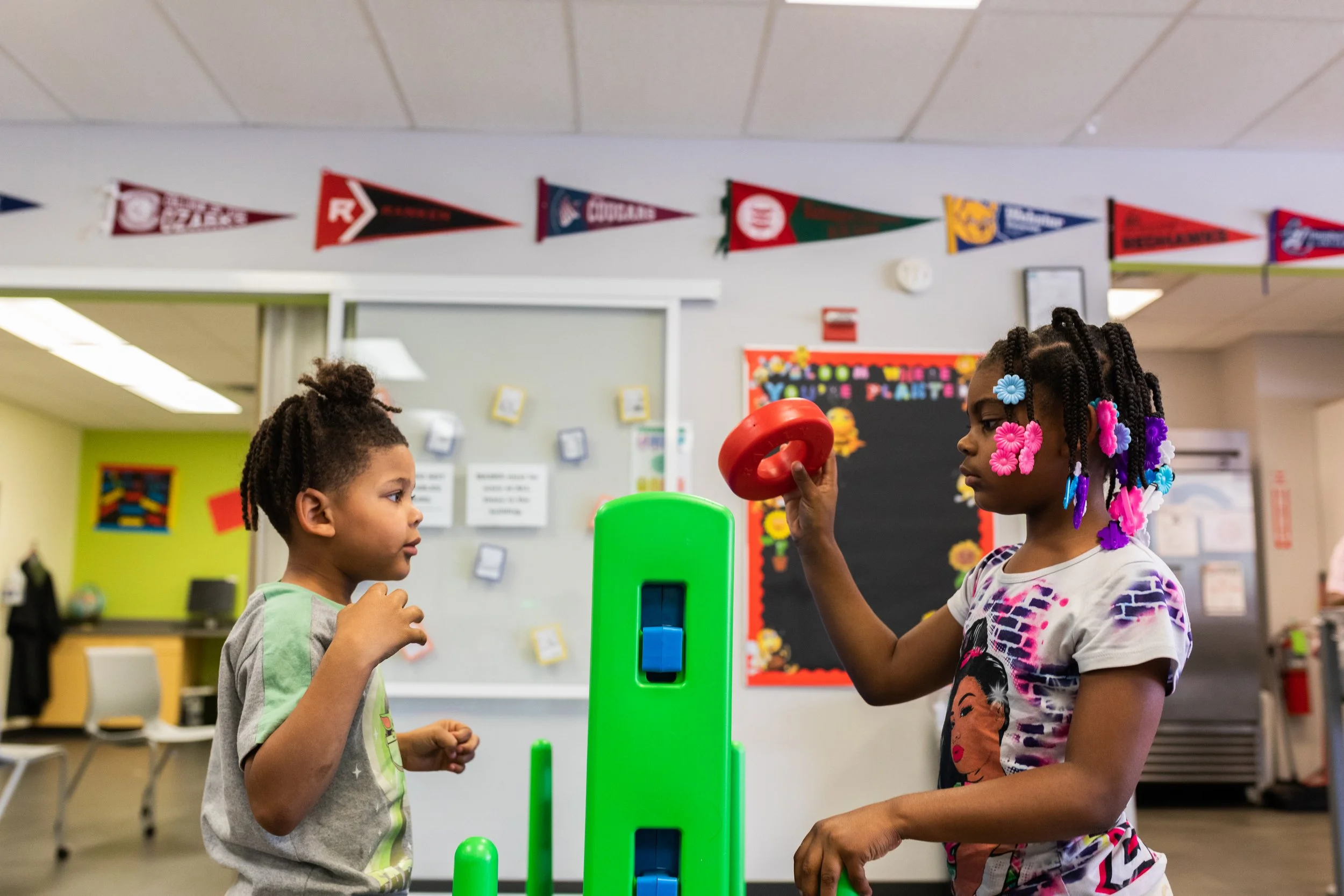 Two young girls are playing with a green marble run and red ring toy in a classroom. One girl with braided hair and colorful hair accessories holds the red ring, while the other girl watches.
