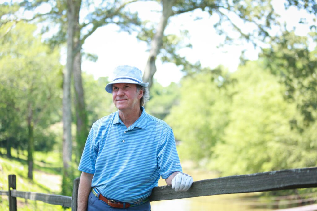 Older man standing by a wooden fence in a park, wearing a blue striped shirt, white gloves, and a light blue hat, with trees and greenery in the background.