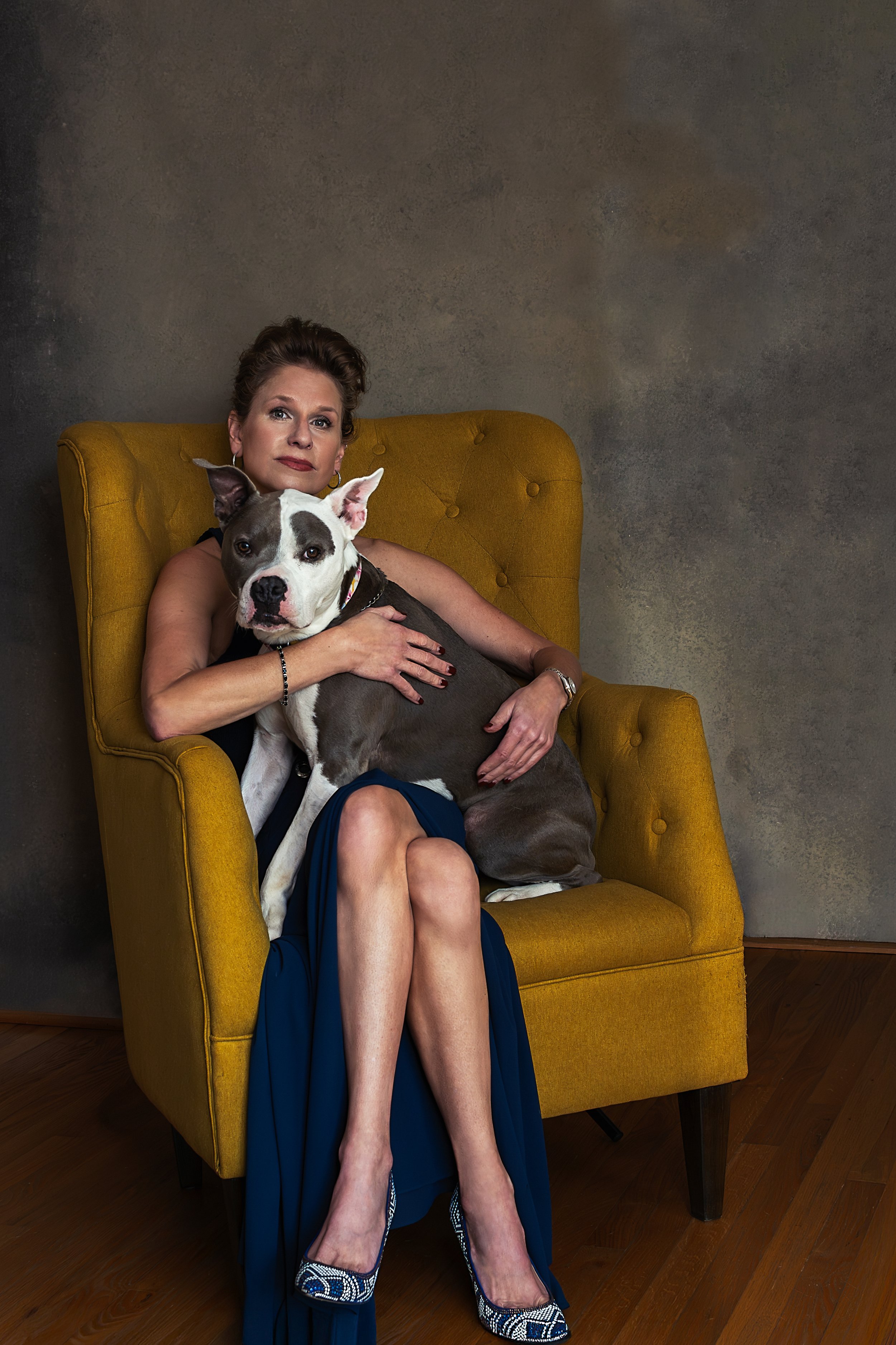 Brunette woman and brown dog with white patches sitting on yellow chair against neutral tone background