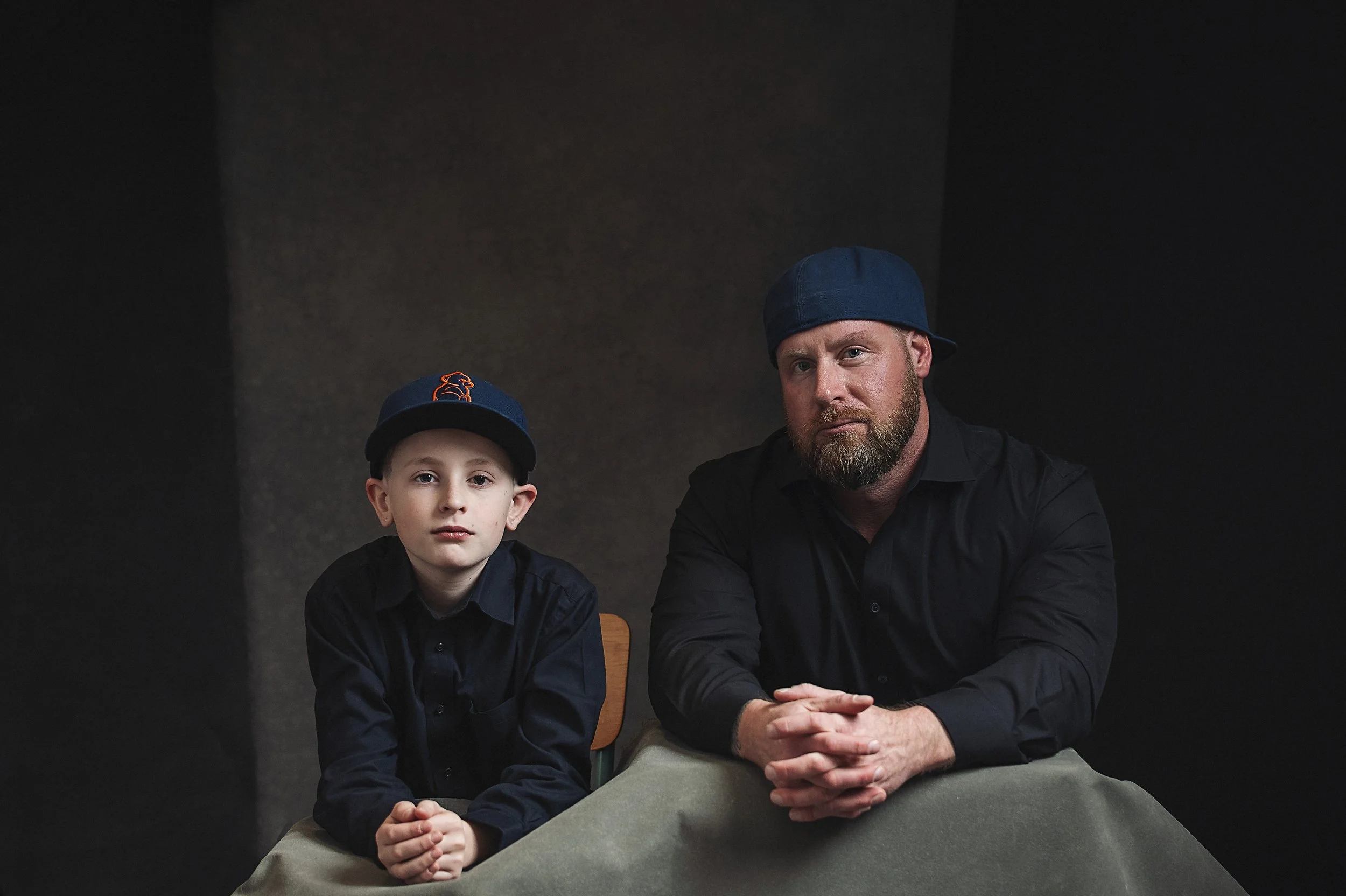 A man and a young boy sitting at a table against a dark background. The boy is wearing a baseball cap with an orange logo and has a serious expression. The man is wearing a blue baseball cap backward and has a beard, with his hand resting on table