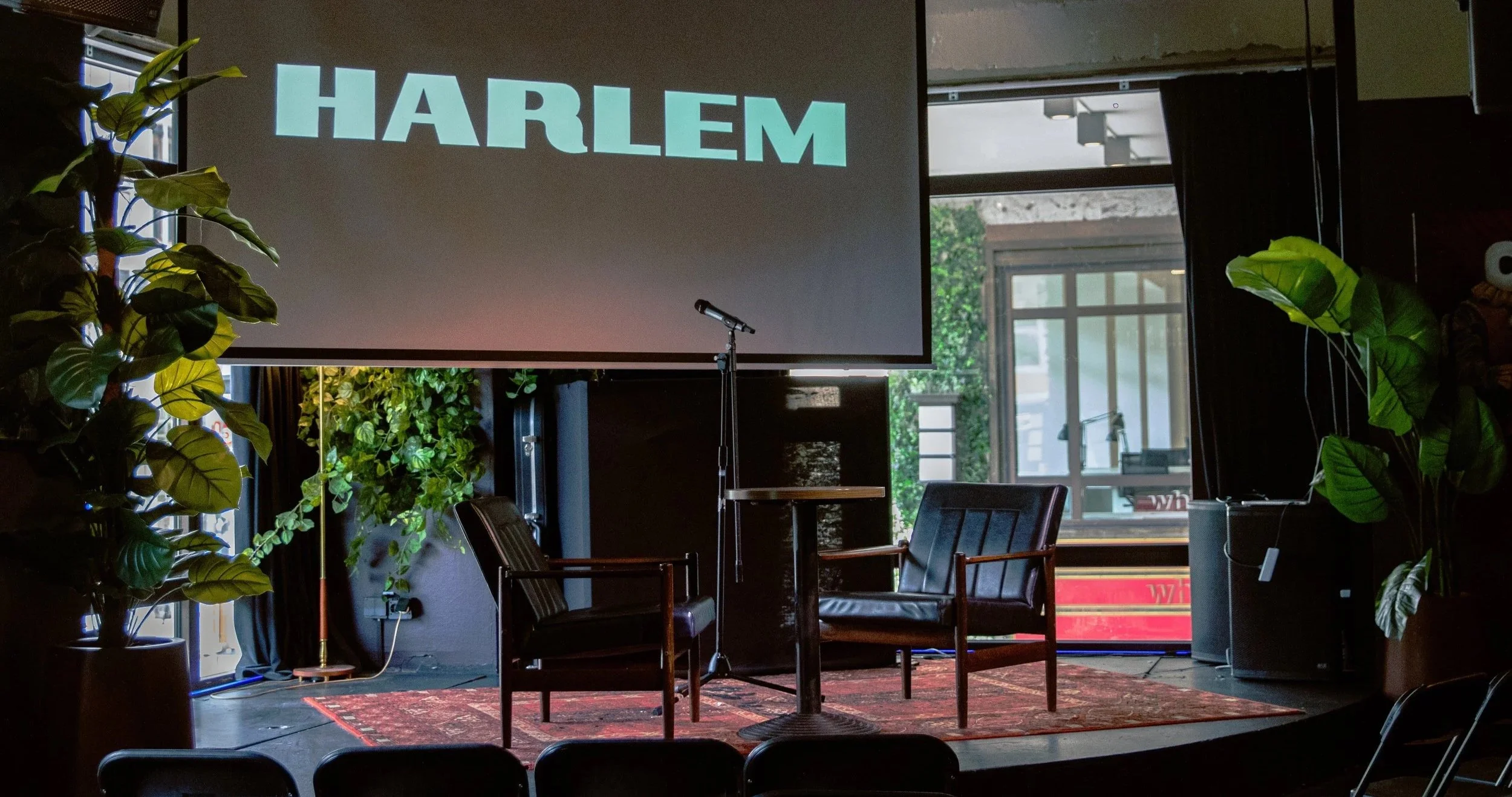 A stage setup featuring a large screen with the word 'HARLEM' projected on it, surrounded by green plants, chairs, a microphone, and a small table, with large windows in the background.