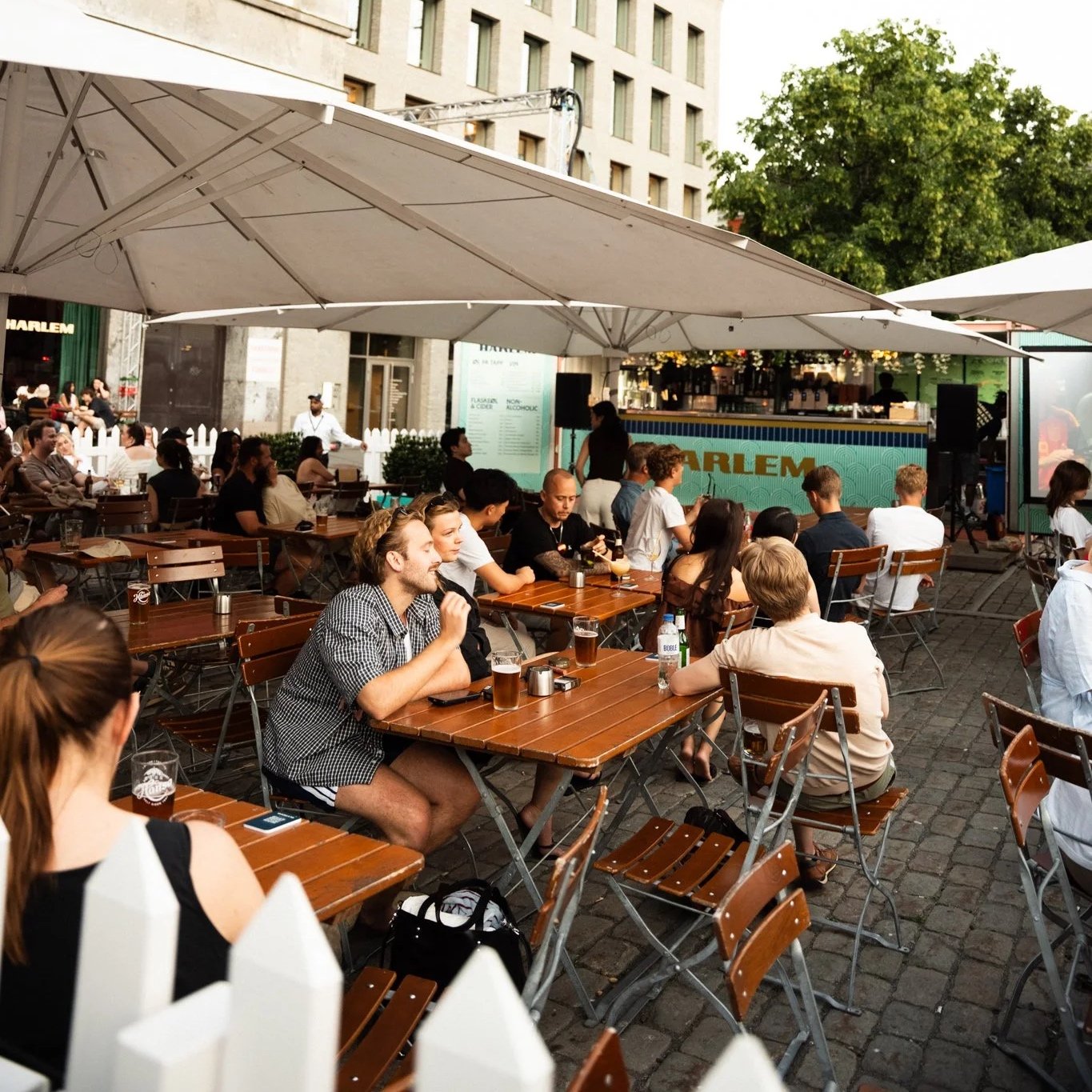 Outdoor patio during daytime with people sitting at wooden tables under large umbrellas, enjoying drinks and socializing, with a bar and a city building in the background.