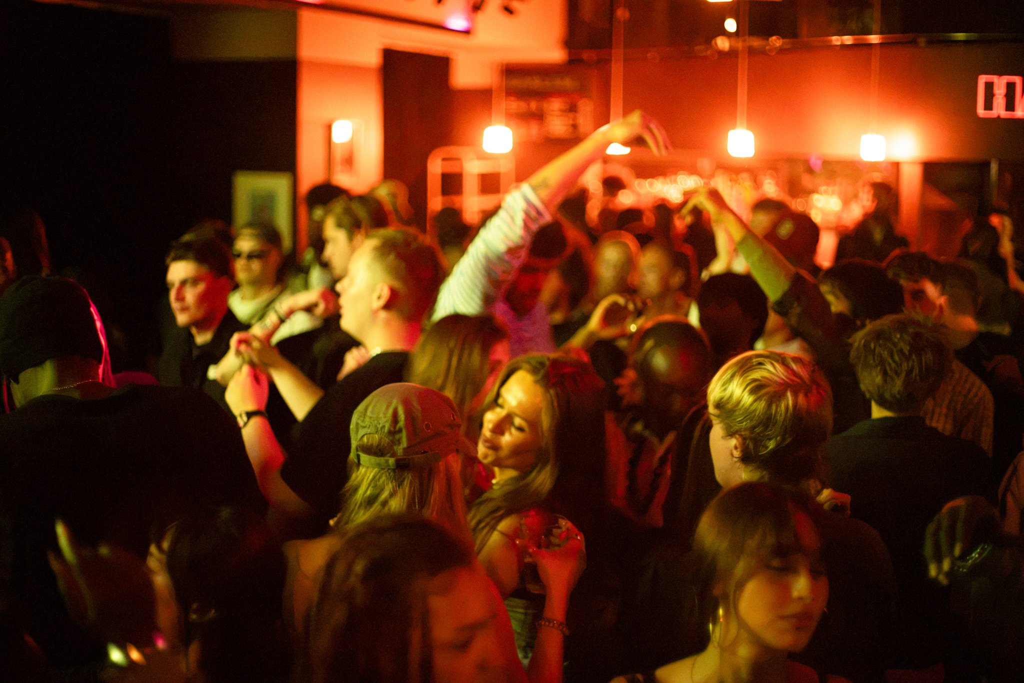 A crowded dance floor in a dimly lit nightclub with red lighting. People are dancing and socializing, with some raising their hands. The background shows bar area and a digital clock display.