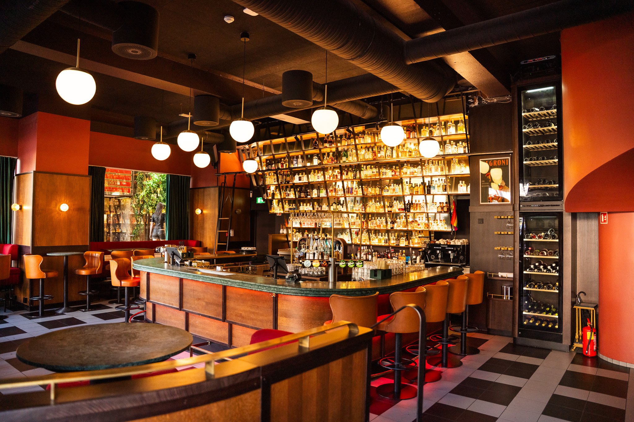 Interior of a modern bar with warm lighting, a curved bar counter, and shelves filled with bottles of alcohol. There are hanging globe lights, orange chairs, and a wine cooler on the right side. A woman can be seen outside through a large window.