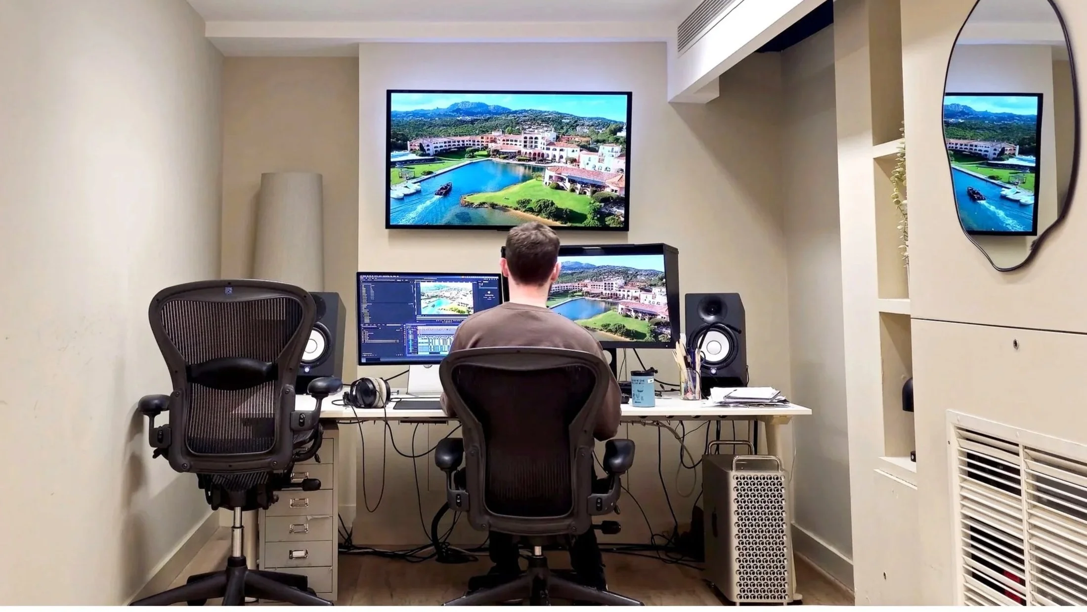 A person working at a desk in a studio or office with two monitors, editing software on one screen, with two large screens displaying an aerial view of a waterfront development and a large mirror on the right wall.