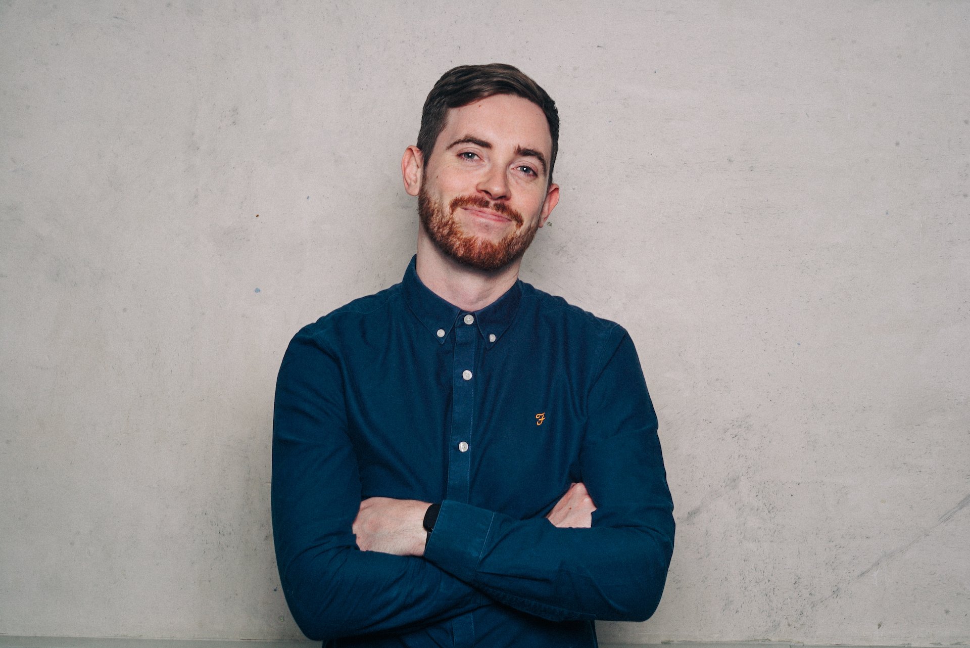 A young man with dark hair, a beard, and light skin, wearing a dark blue button-down shirt with crossed arms, standing against a plain light-colored wall, smiling slightly.