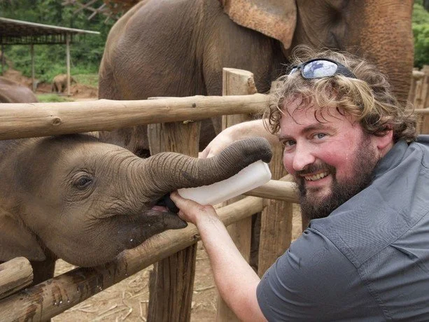 A man feeding a young elephant with a bottle at a zoo or sanctuary, with larger elephants in the background.