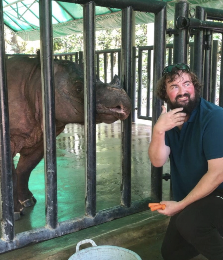 A man with glasses and curly hair, kneeling next to a hippopotamus behind metal bars in a zoo or animal sanctuary cage, holding carrot sticks.