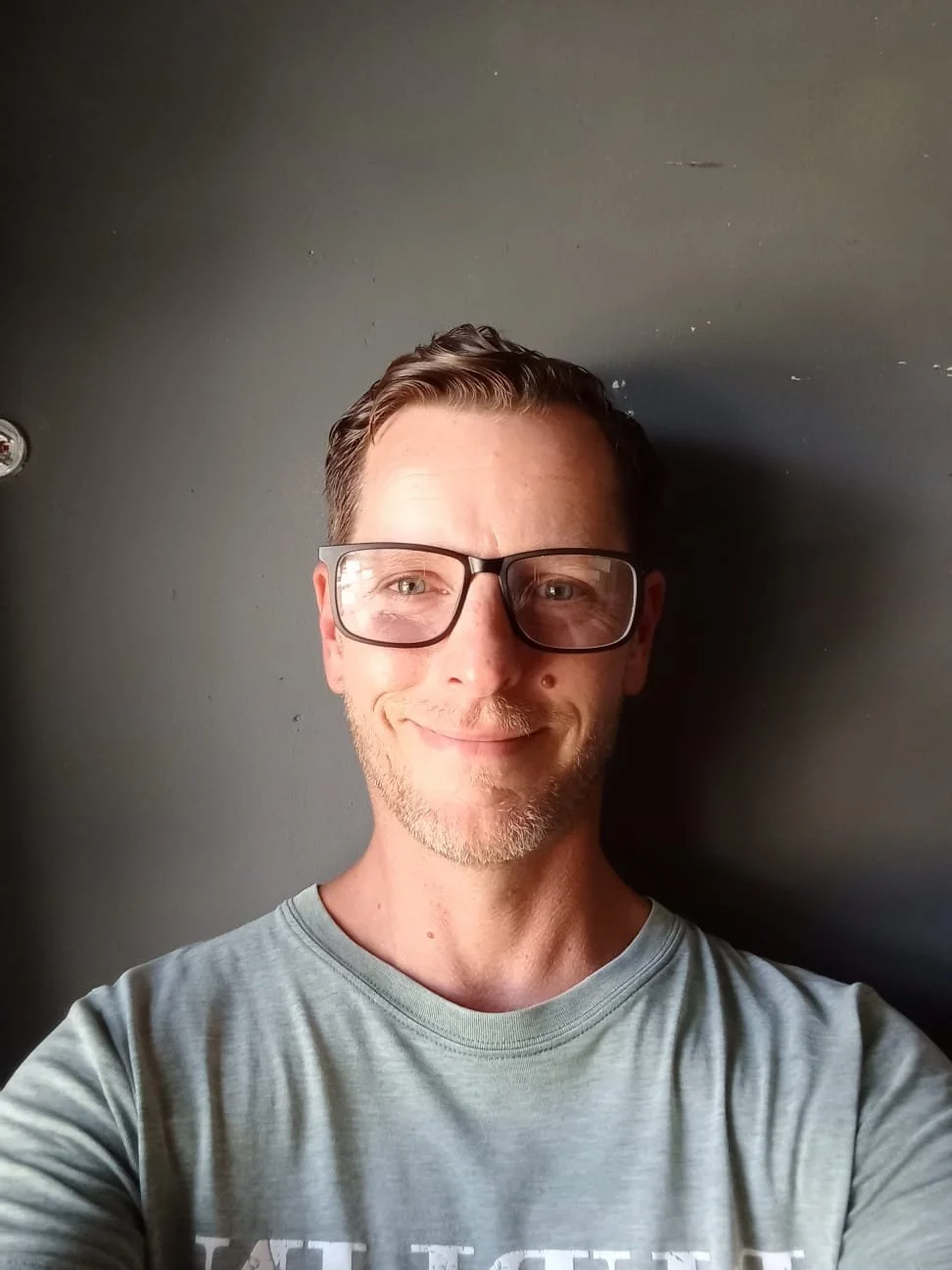 A man with glasses and a light beard taking a selfie against a dark wall. He is smiling and wearing a gray T-shirt.