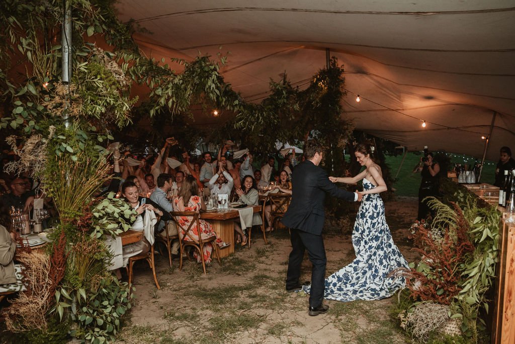 A bride and groom dancing at their wedding reception under a decorated outdoor tent with guests sitting at tables watching and cheering.