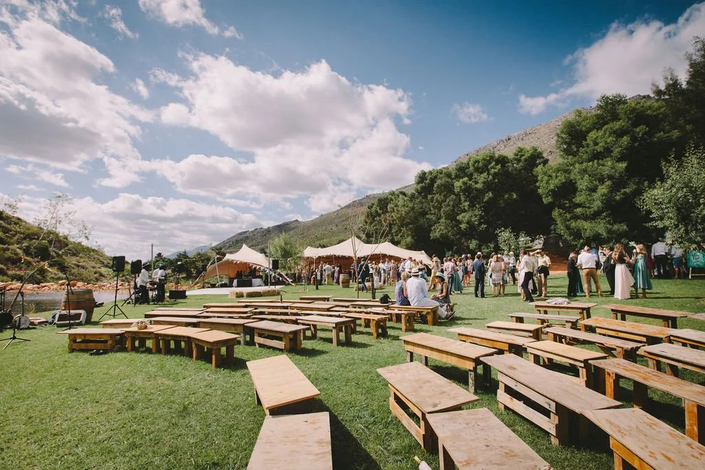 Outdoor wedding reception setup with benches and tents, guests socializing, in a mountainous landscape with blue sky and white clouds