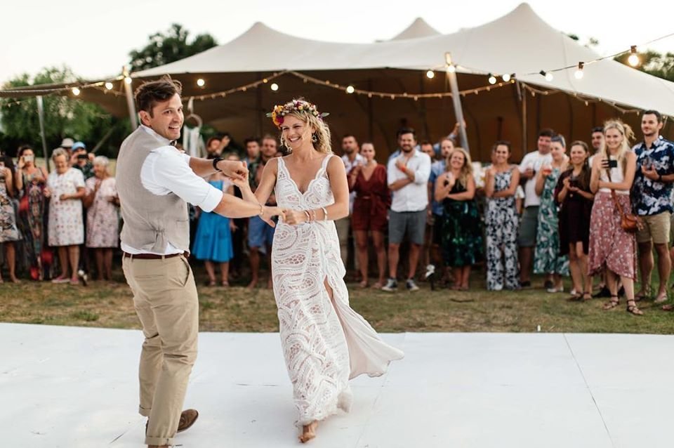 Couple dancing at an outdoor wedding reception with guests watching, under string lights and a large tent in the background.