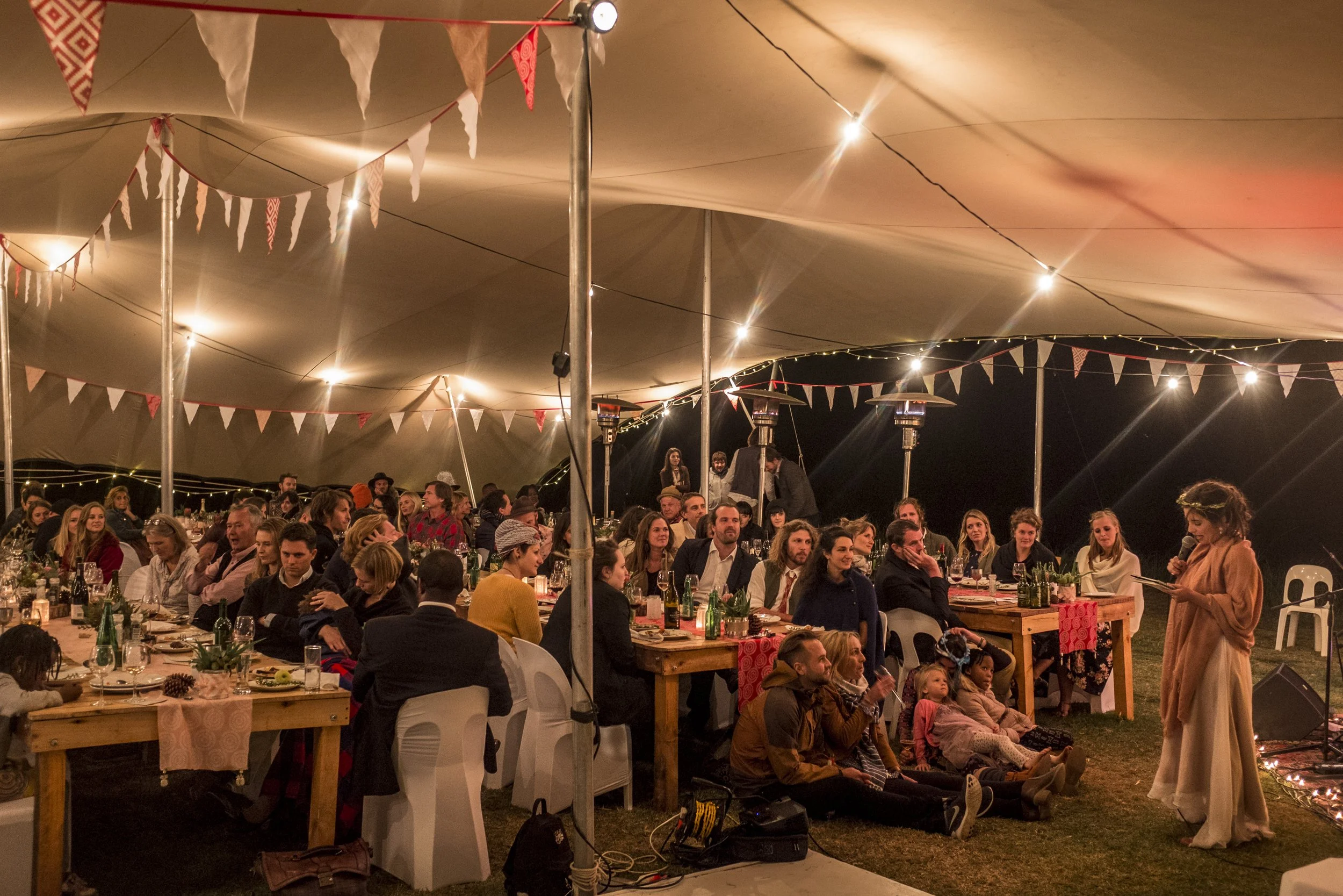 People attending an outdoor event under a large tent decorated with string lights and triangular flags, with a woman speaking into a microphone on the right side.