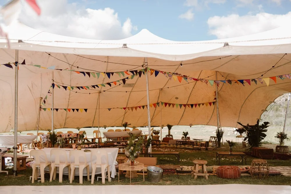 Decorative outdoor event tent with colorful bunting, wooden tables, white chairs, and floral centerpieces under a blue sky.