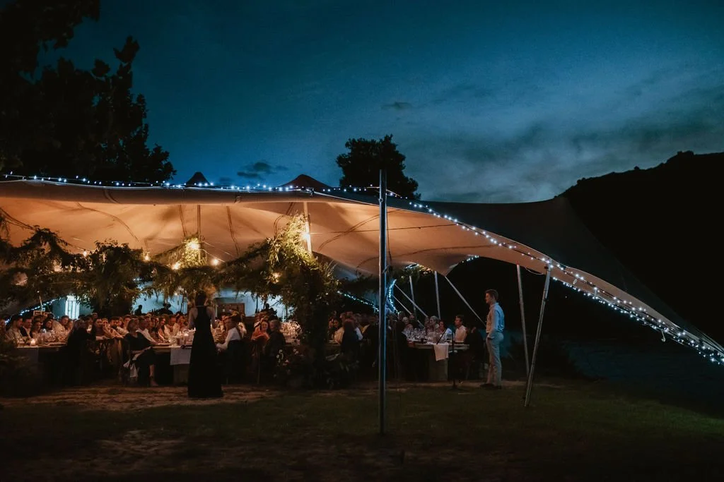 An outdoor event or celebration takes place under a large tent with string lights at night. Guests are seated at tables, and people are standing or walking around. The scene is set against a dark sky with silhouettes of trees and hills.