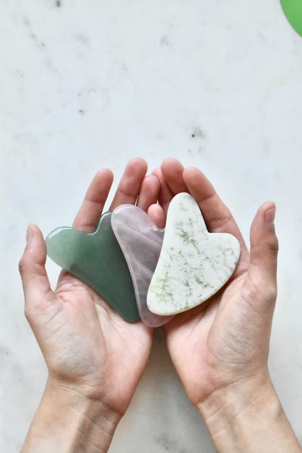 Hands holding three polished stones in the shape of hearts, in shades of green, gray, and white with green veining, on a light gray surface.