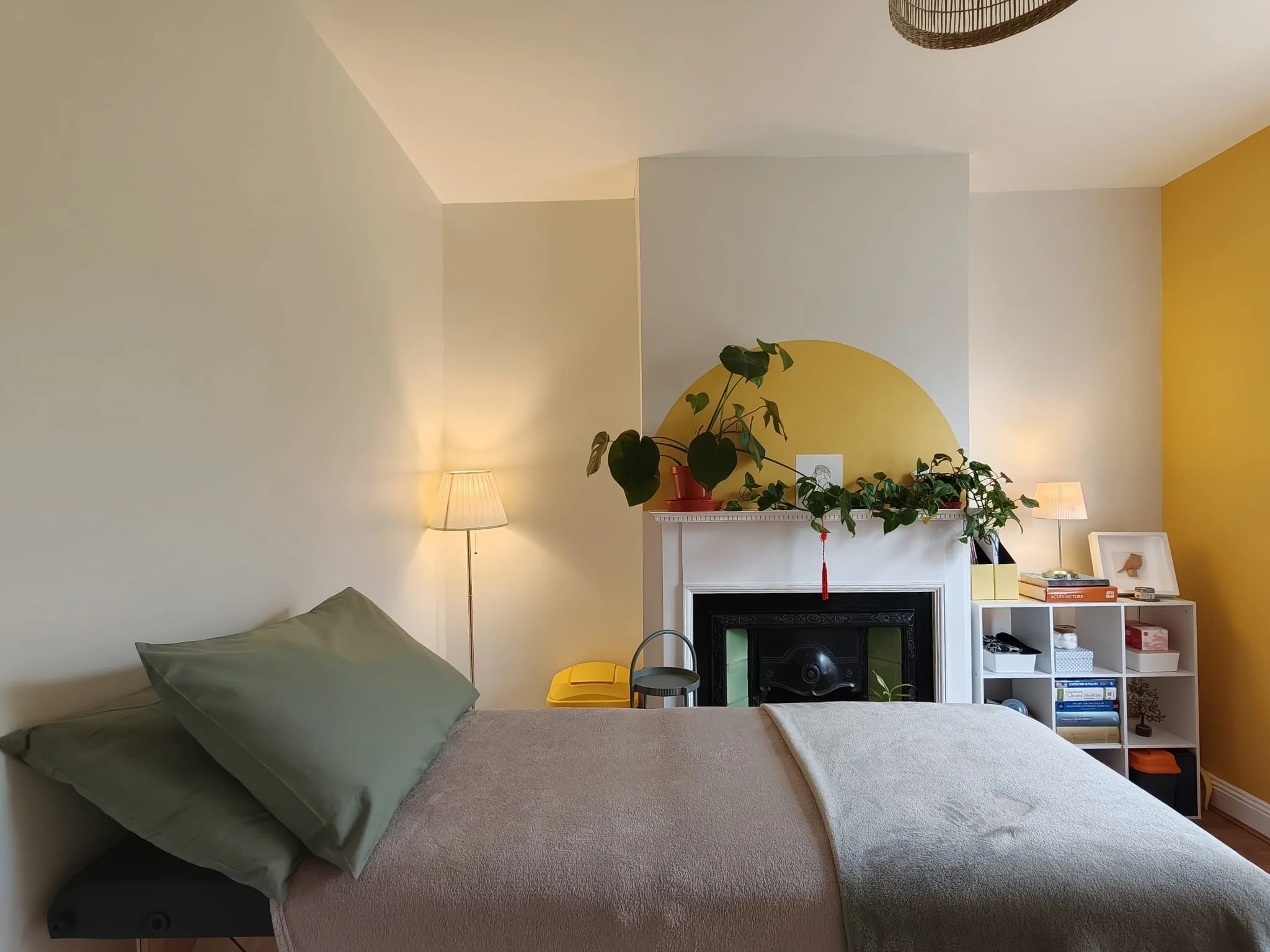 A bedroom with a bed, green pillow, and beige blanket; two lamps on bedside tables; a white mantel above a black fireplace with green trim; potted plant and artwork on the mantel; white shelving unit with books and decor; yellow accent wall.