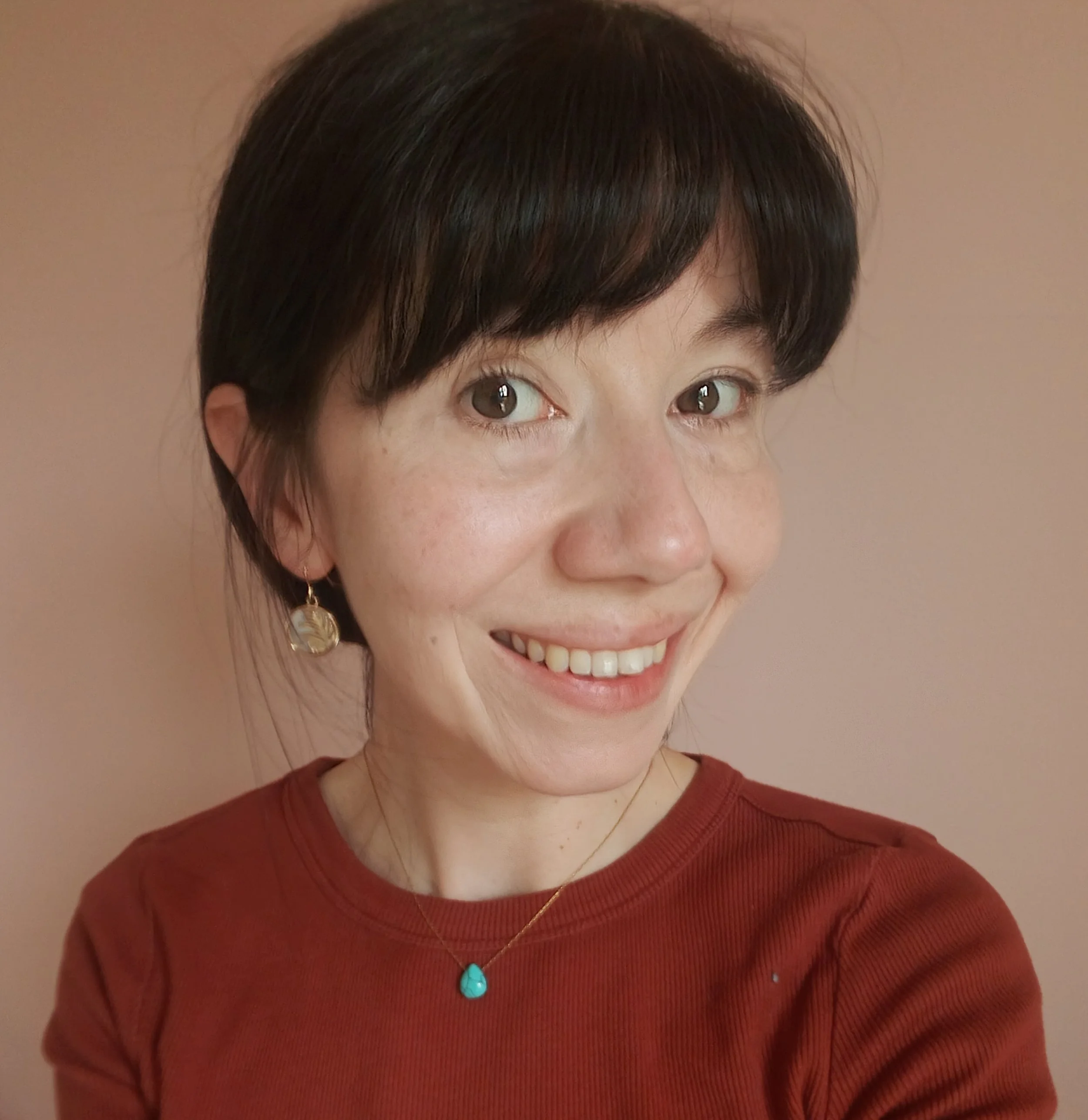 Close-up of a smiling woman with short dark hair, wearing earrings, a turquoise necklace, and a red shirt, against a pink background.