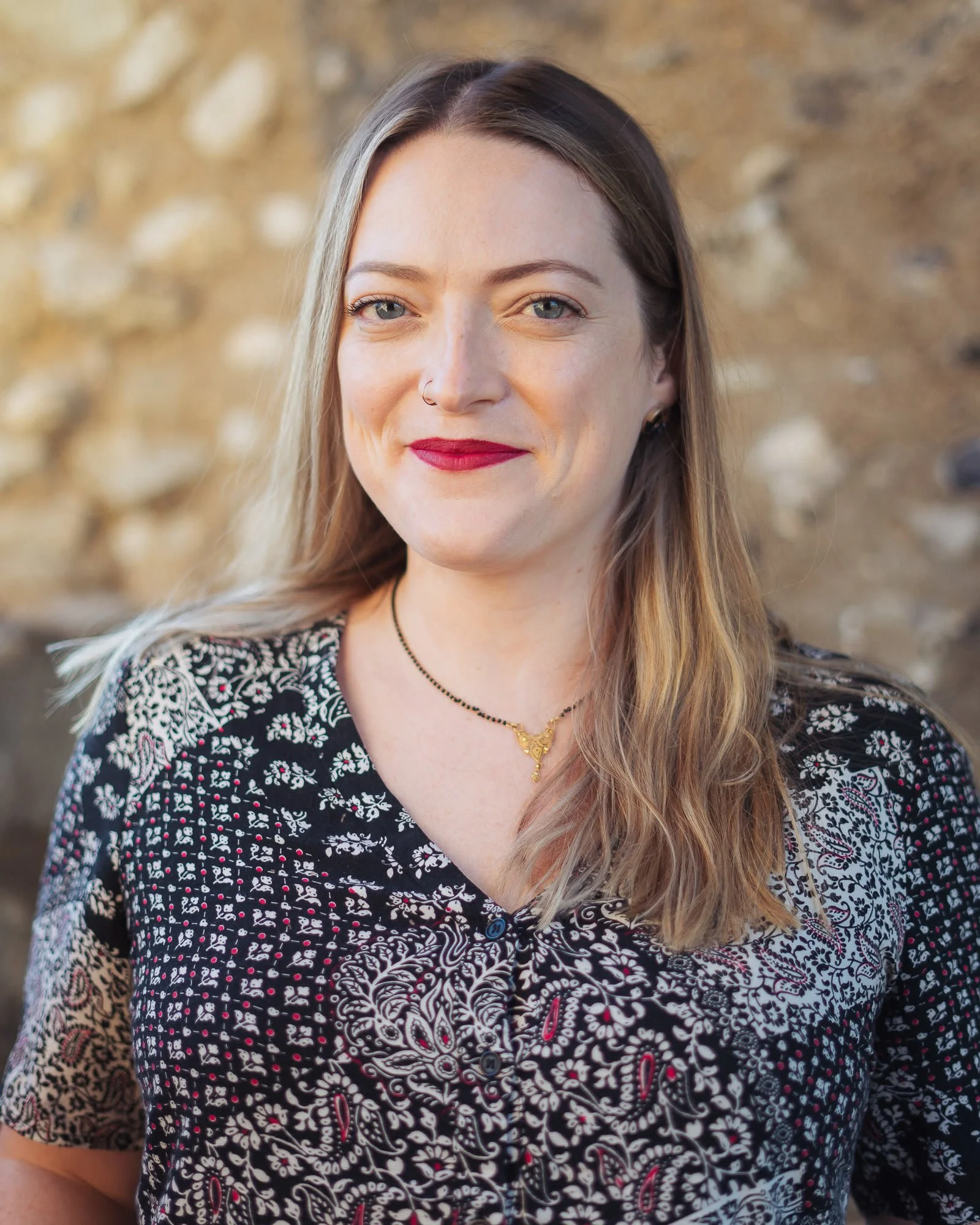 A woman with long brown hair, blue eyes, and a nose ring, smiling outdoors with a stone wall in the background, wearing a black and white patterned shirt and a gold necklace.