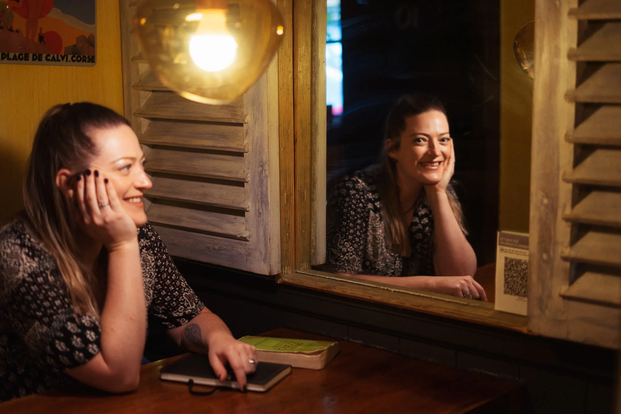 A woman sitting at a table, looking into a mirror, smiling. The mirror reflects her face and a wooden louvered window nearby. There is a notebook and a book on the table.