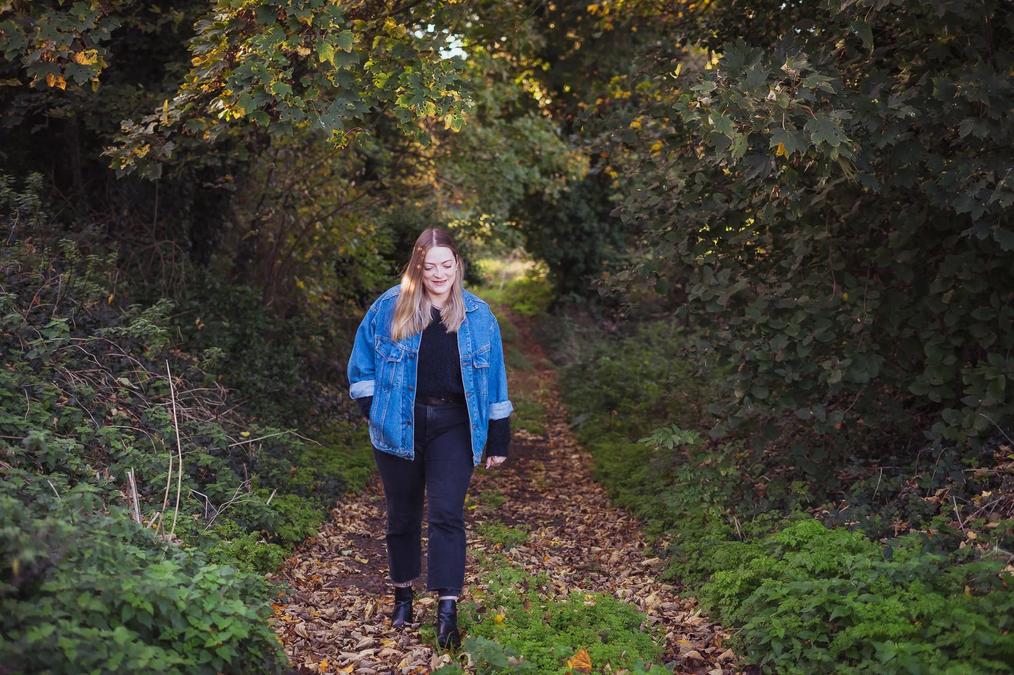 A woman with blonde hair wearing a denim jacket, black top, black pants, and black ankle boots walking along a leaf-covered trail through a green forest in autumn.