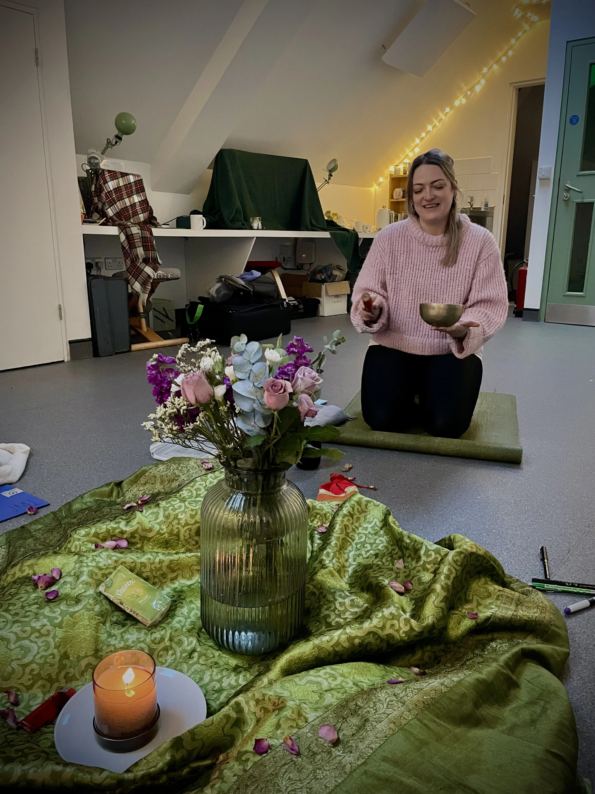 A woman kneeling on a green mat in a room decorated with fairy lights, arranging flowers in a vase on a green patterned fabric, with a lit candle nearby. She is wearing a pink knitted sweater and has long hair.