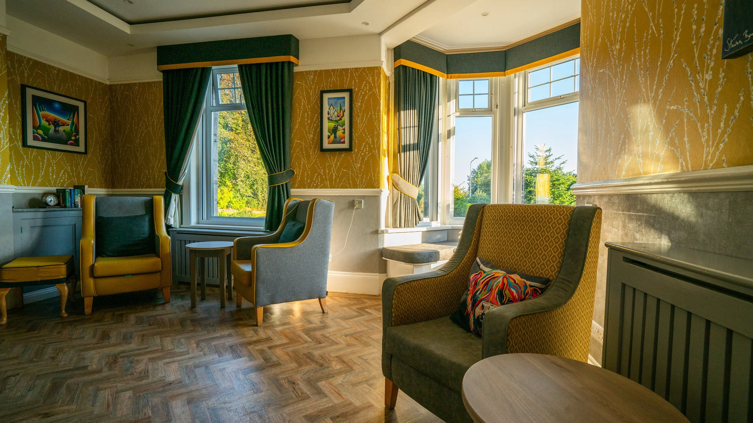 A cozy living room with large windows letting in natural sunlight. There are colorful chairs, a small table, and artwork on the walls. Yellow and green curtains frame the windows, and the room has a wooden floor.