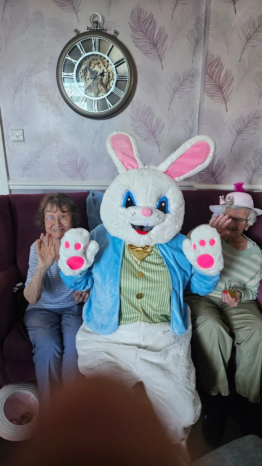 Two elderly women sitting on a purple couch with a person dressed as a large white bunny in the middle. The woman on the left is wearing glasses, a striped shirt, and is waving. The woman on the right is wearing glasses, a hat with a pink decoration, and holding a glass. The bunny costume has large pink and white paws, big blue eyes, pink nose, and is wearing a blue jacket and yellow striped shirt.