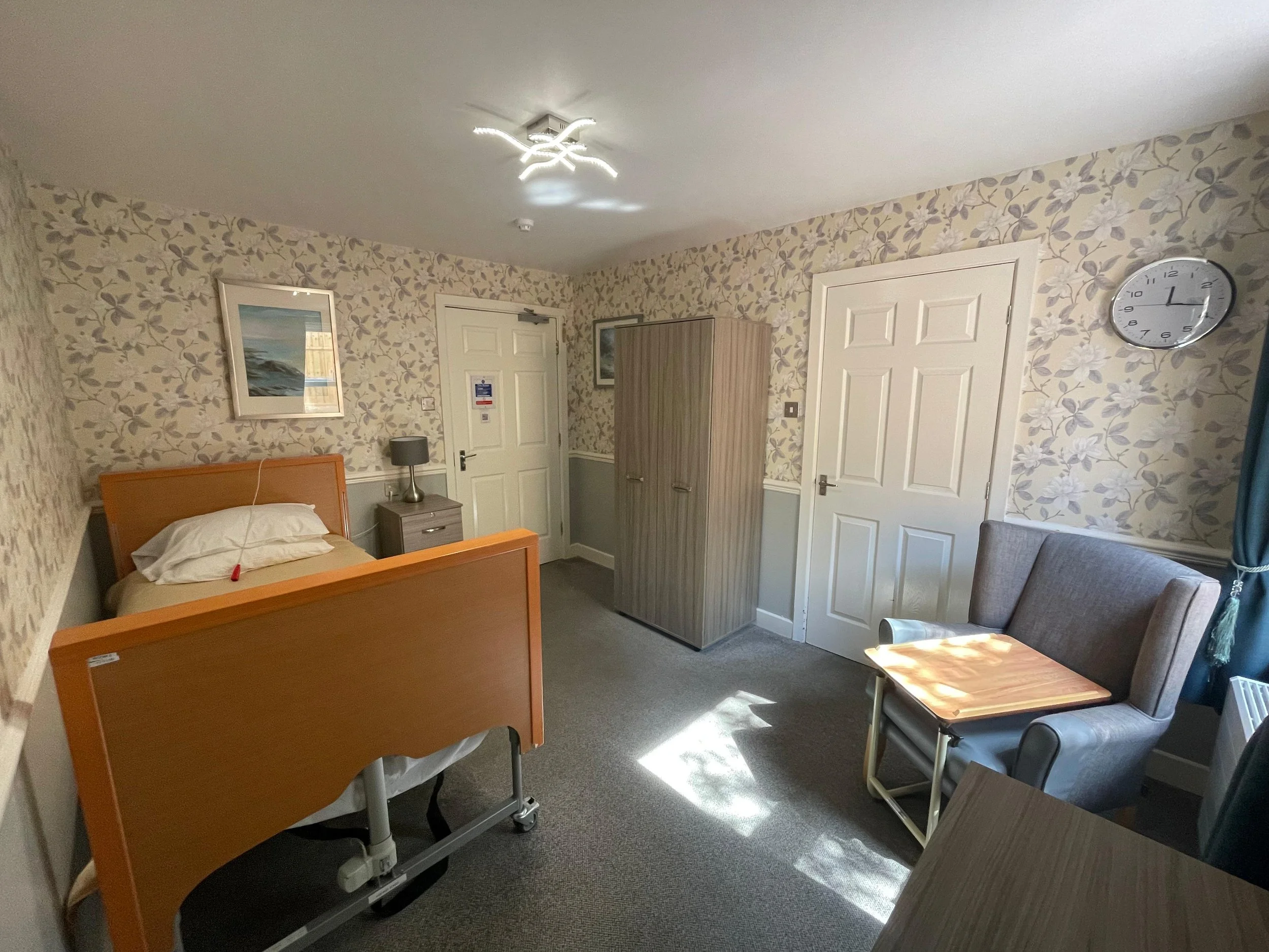 A hospital room with floral wallpaper, a hospital bed with white sheets, a bedside table with a lamp, a wardrobe, a gray armchair, a small wooden table, a clock on the wall, and a ceiling light.