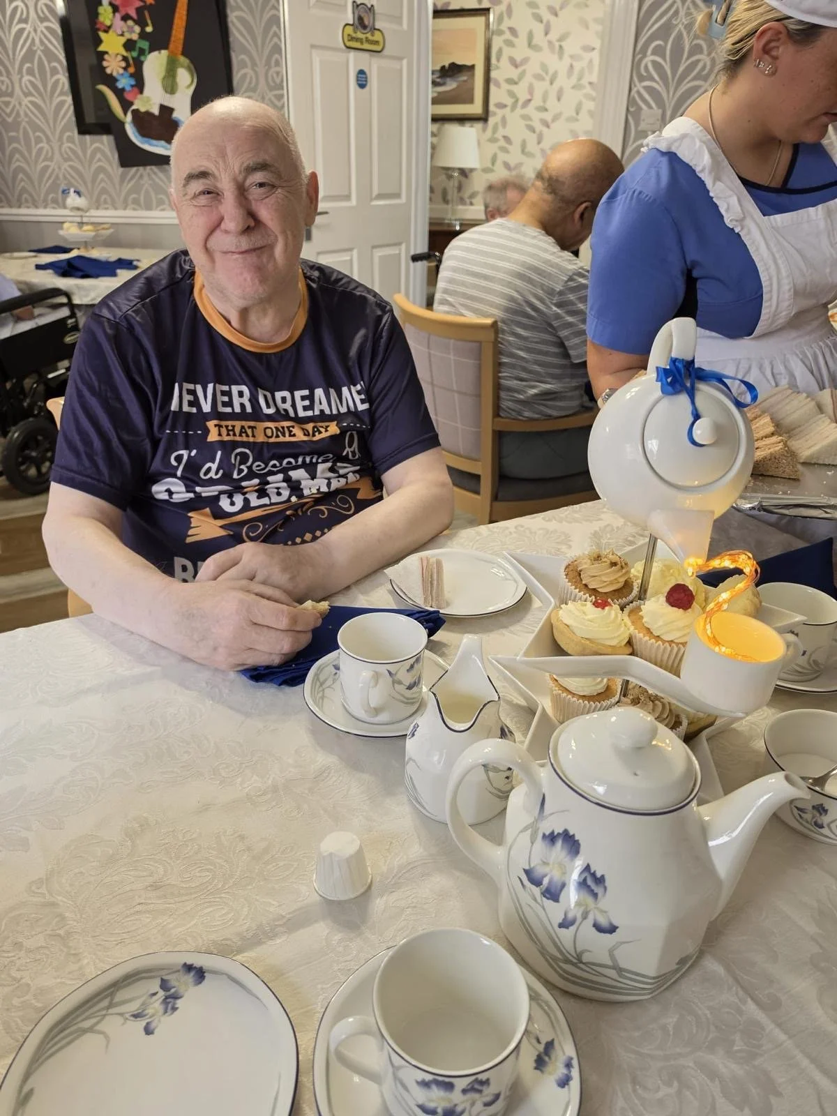 An elderly man sitting at a table with a tea set and a tiered tray of cupcakes, smiling at the camera. A woman in a blue uniform is nearby, and there are other people in the background in a decorated room, possibly a shared care or nursing home setting.