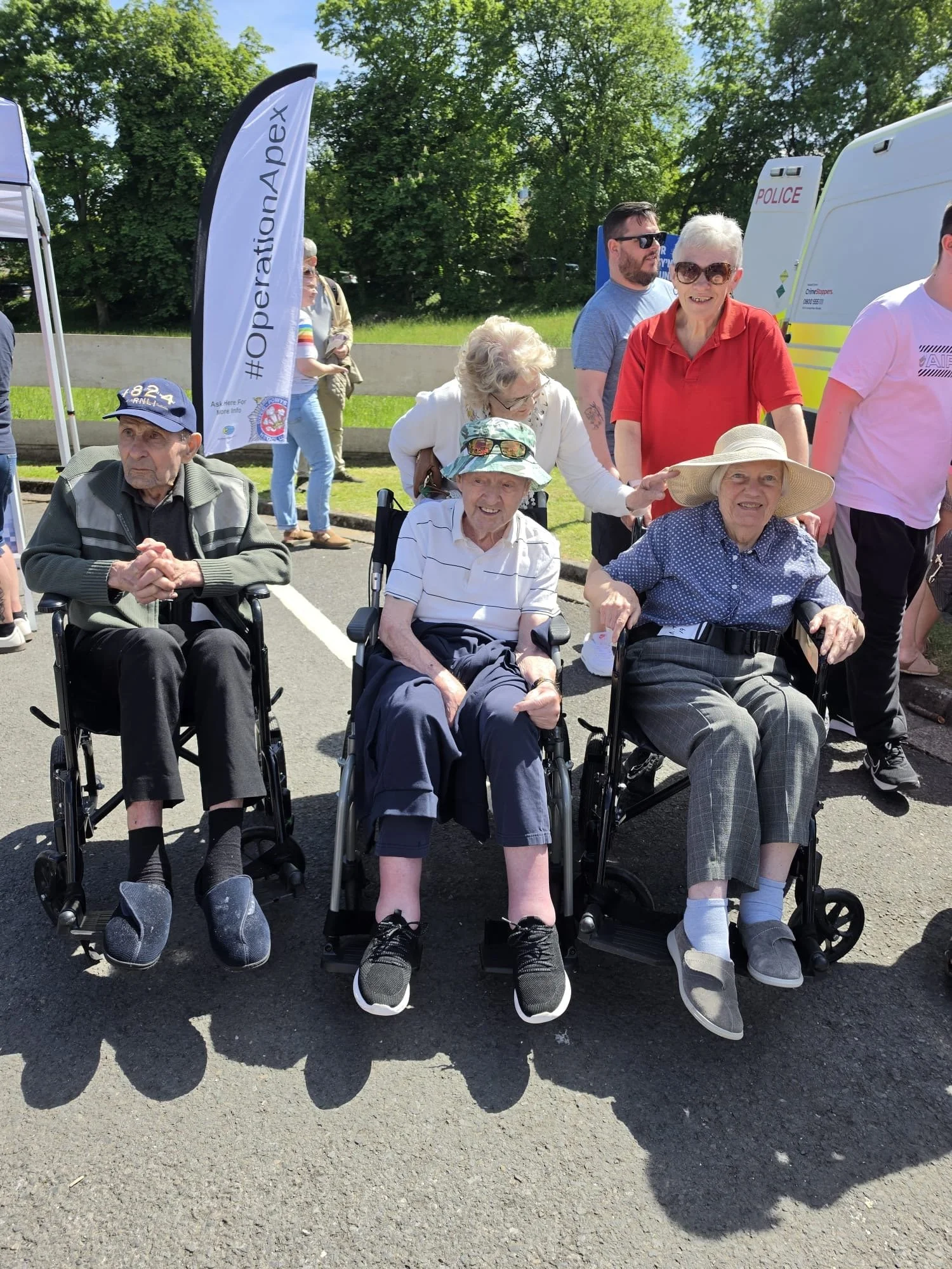 Group of elderly and middle-aged people, some in wheelchairs, outdoors at a sunny event with trees in the background. Several people are smiling and interacting.