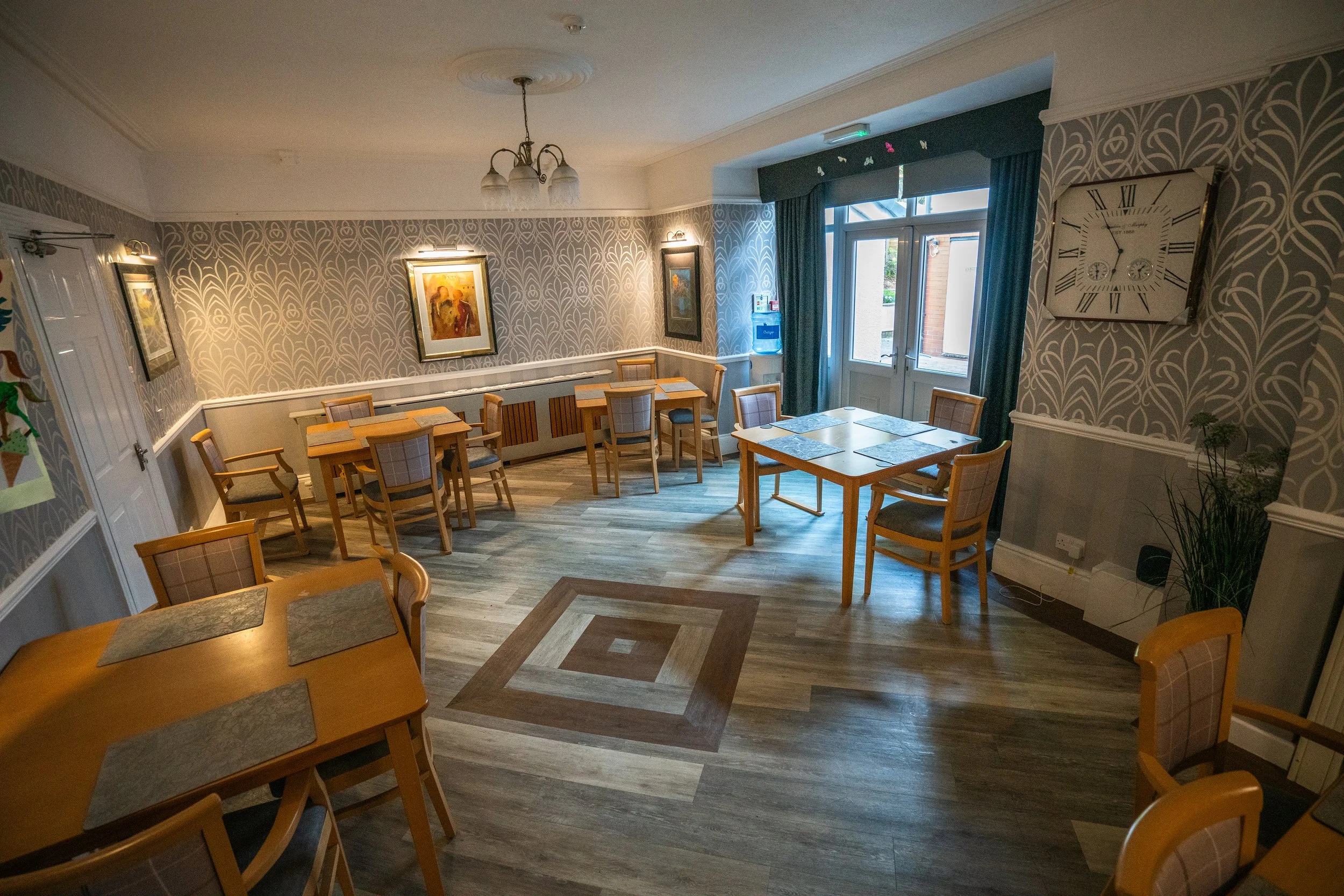 A cozy dining area with wooden tables and chairs, patterned wallpaper, framed artwork, and large windows with blue curtains, illuminated by a ceiling chandelier and wall lights.