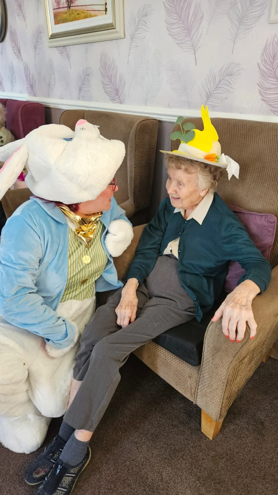 An elderly woman wearing a festive Easter hat with a yellow bunny and green leaves, sitting on a beige armchair, conversing with a person in an Easter Bunny costume in an indoor setting with purple feather-patterned wallpaper.
