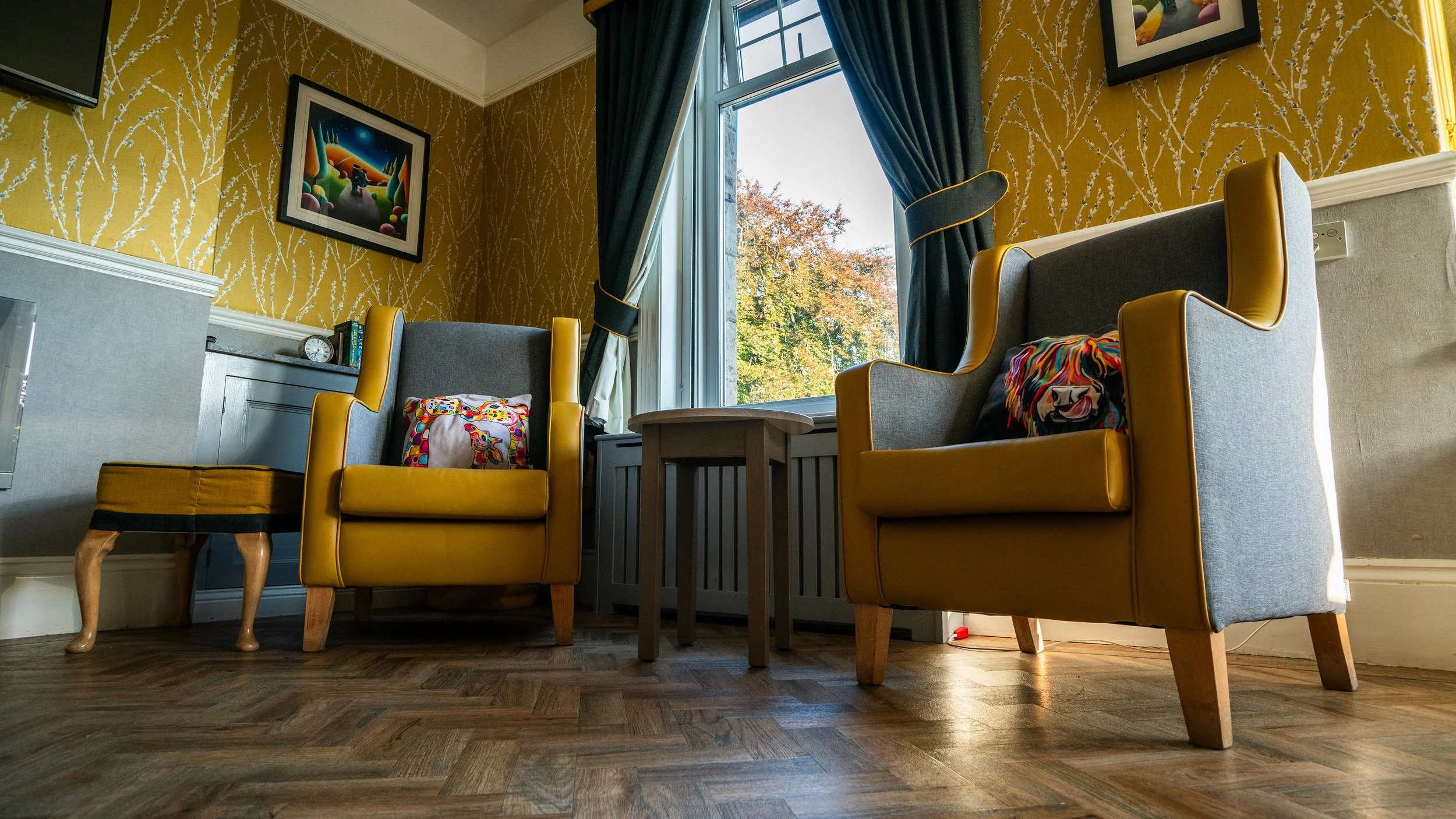 A cozy sitting area with two mustard yellow armchairs, a small wooden stool, a framed colorful picture on the wall, a window with blue curtains, and a view of trees outside.