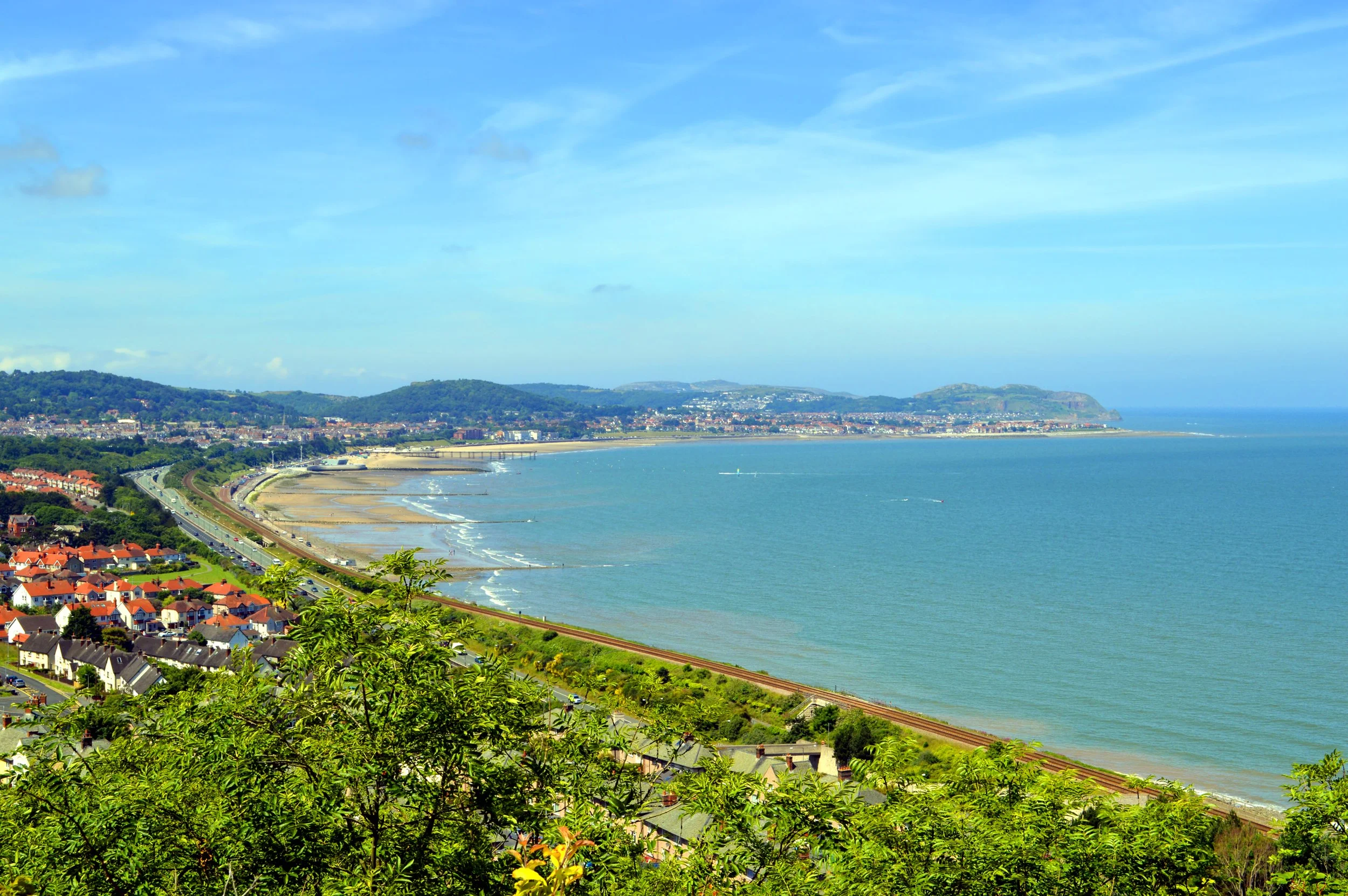 A coastal landscape view showing a beach, a town with houses, and green hills under a partly cloudy sky.