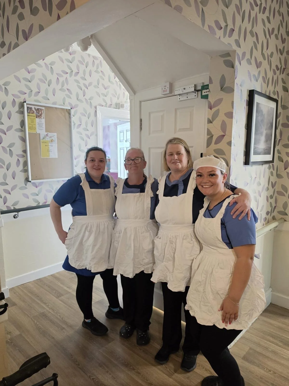 Four women wearing uniforms and aprons standing together inside a cafe or restaurant, smiling at the camera.