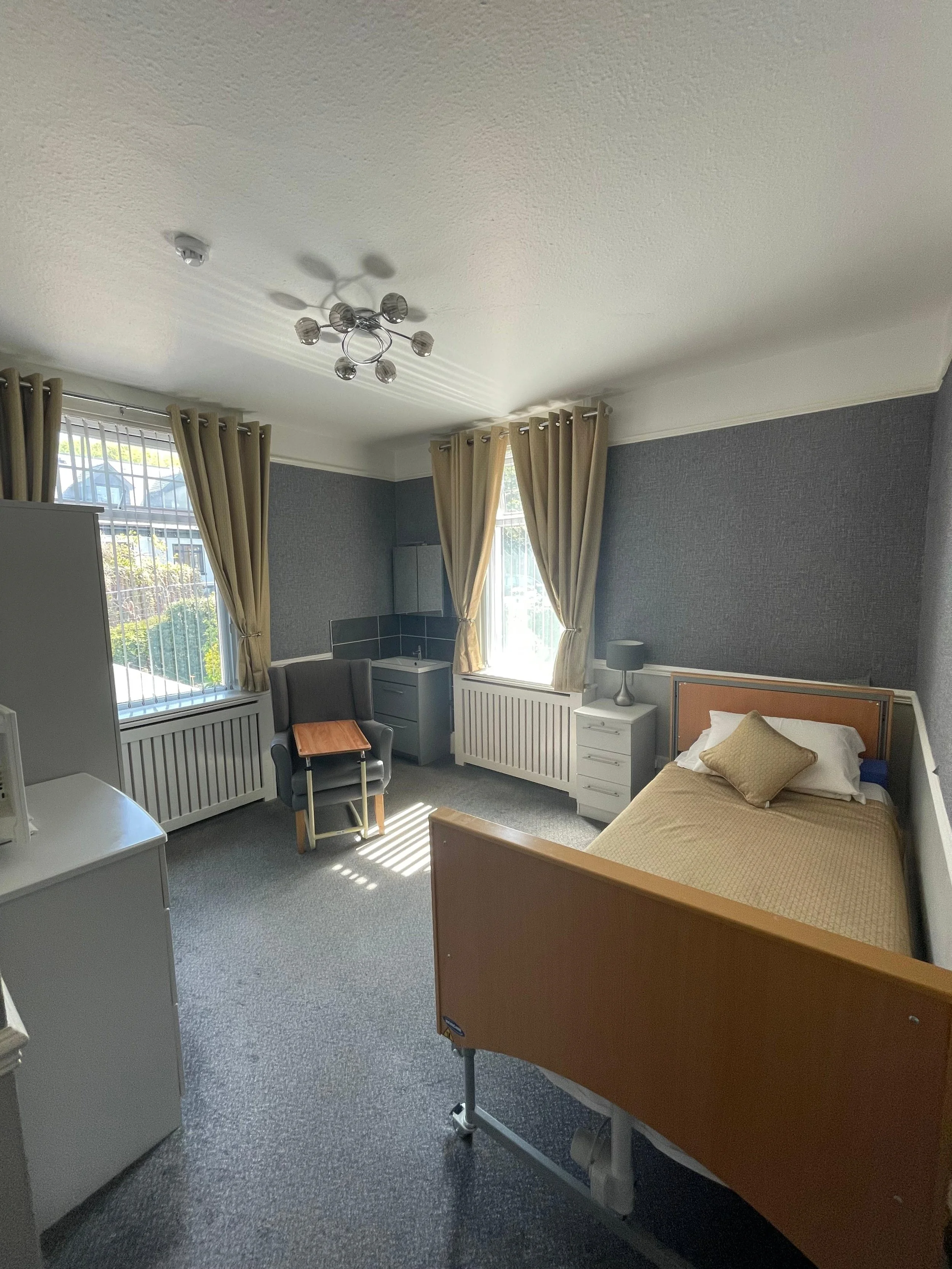 Hospital room with a bed, a gray armchair with a small wooden table, two large windows with beige curtains, gray walls, and a white nightstand with a gray lamp on it.