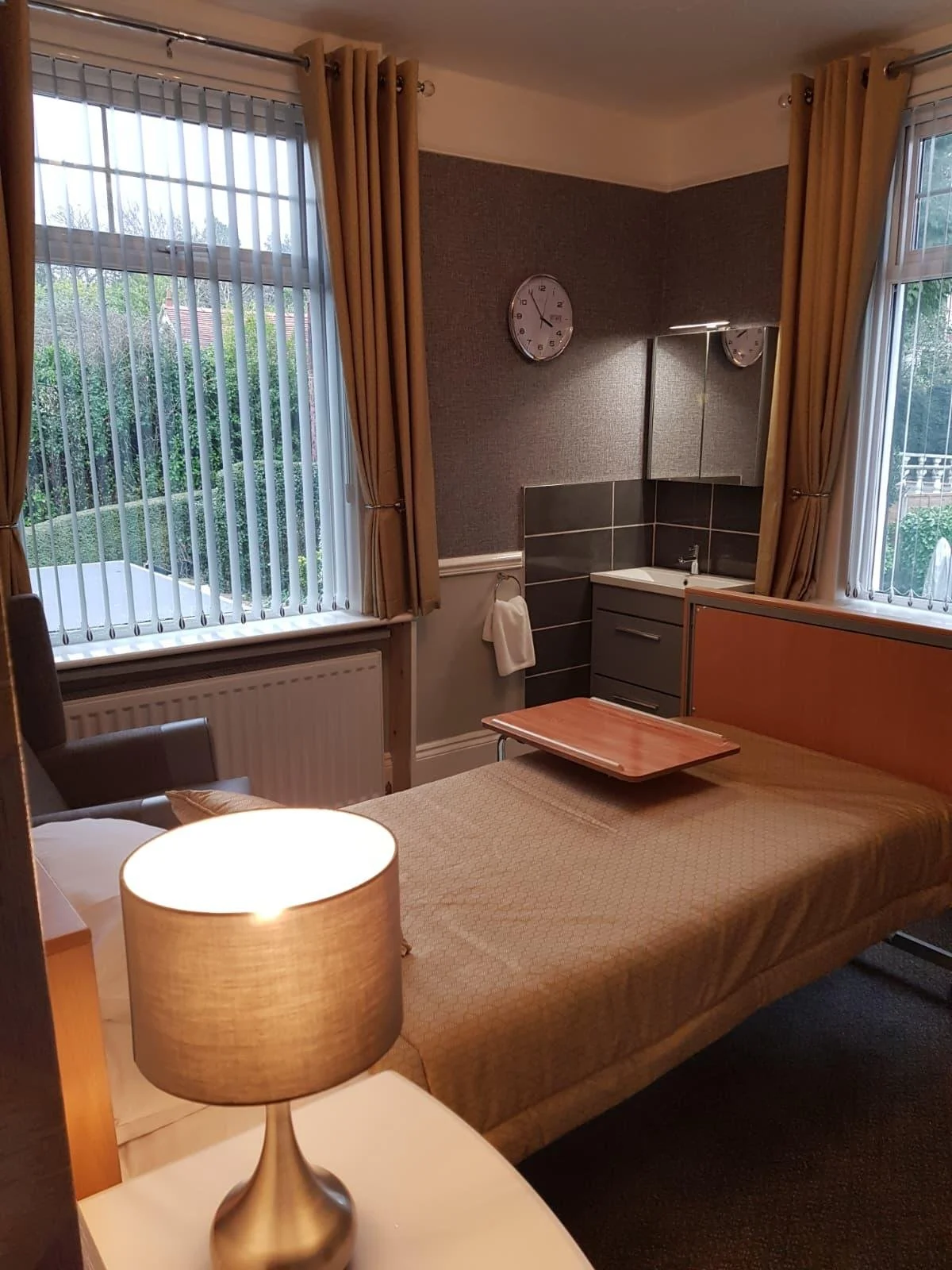 A hotel room with two large windows with beige curtains, a bed with a bedside lamp, a small desk with a tray, a wall clock, and a mini sink area.