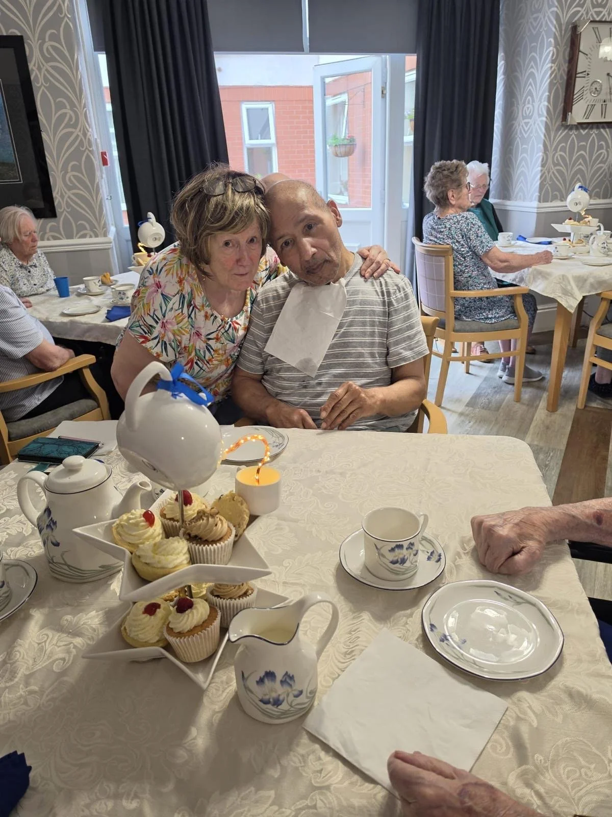 Elderly woman and man with a bib celebrating at a tea party with cupcakes, tea cups, and a tiered cupcake stand in a decorated dining room.