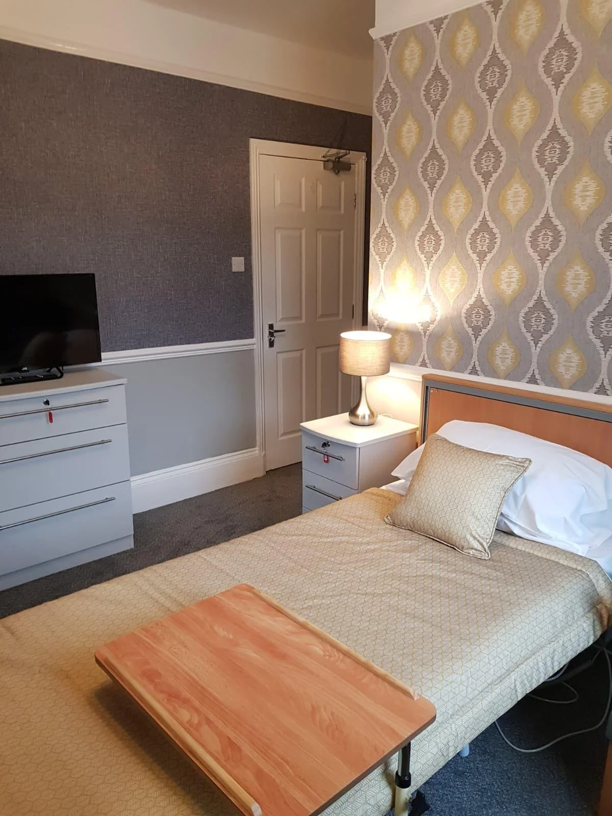 A hotel room with a bed, bedside table, lamp, TV, and a dresser. The room features patterned wallpaper and grey wall paint.