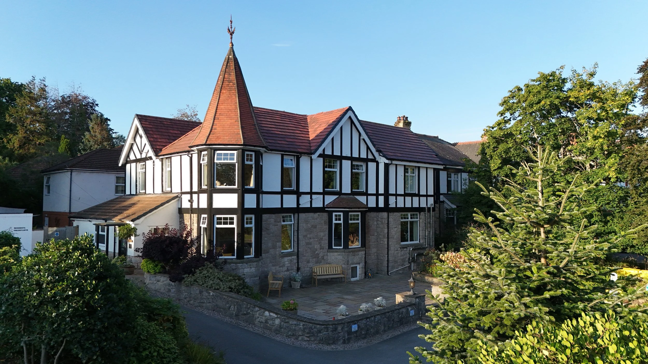 A large house with black and white Tudor-style architecture, stone foundation, multiple windows, and a steeply pitched roof with a tall, pointed turret. Surrounded by trees and landscaped garden.