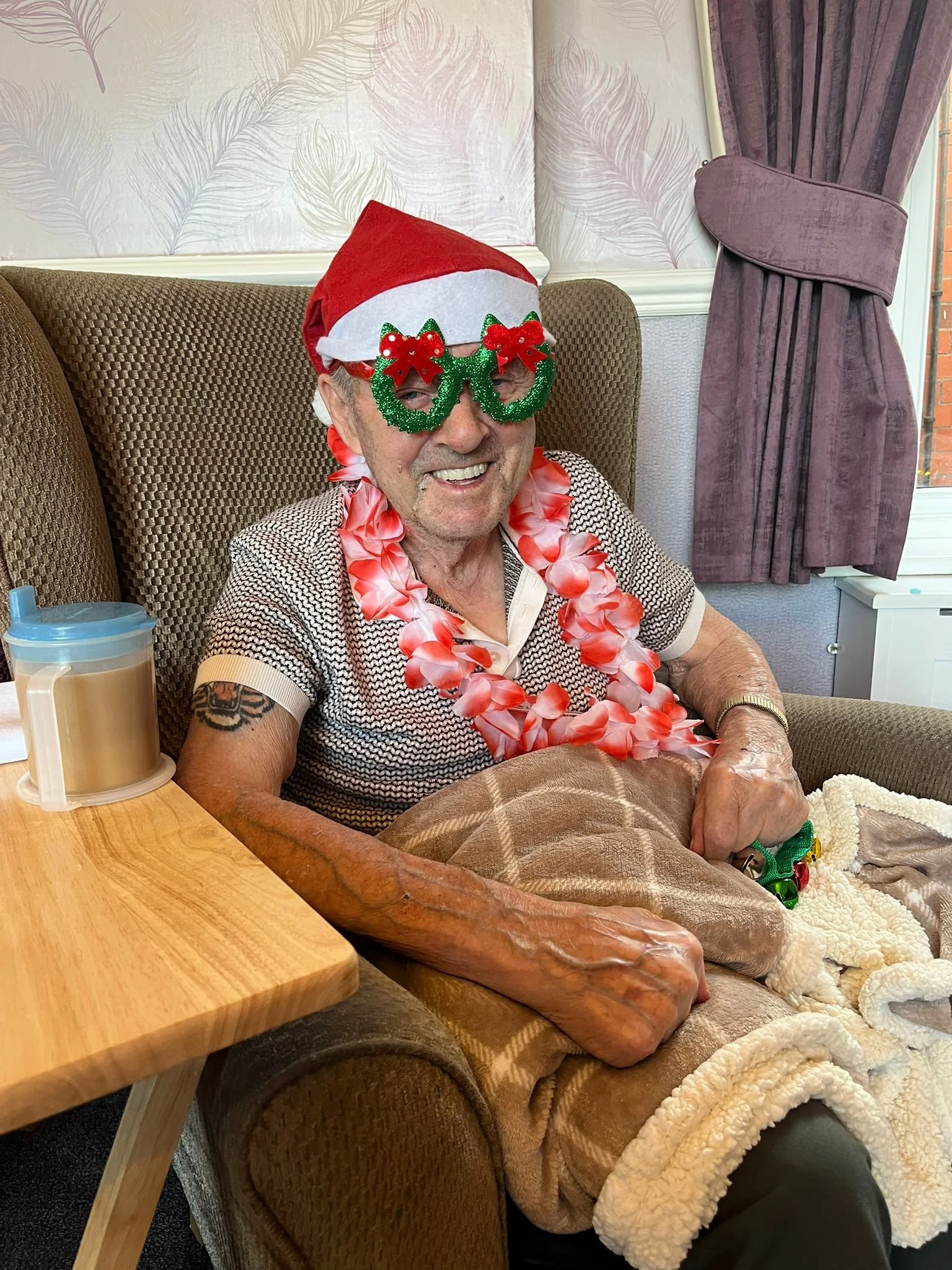 A smiling elderly man sitting in a chair, wearing holiday-themed accessories including green glittery glasses with red bows, a Santa hat, and a pink Hawaiian lei. He has a towel over his lap and a blanket covering his legs. There is a table with a cup to his right, with curtains and a wall with fern-patterned wallpaper in the background.