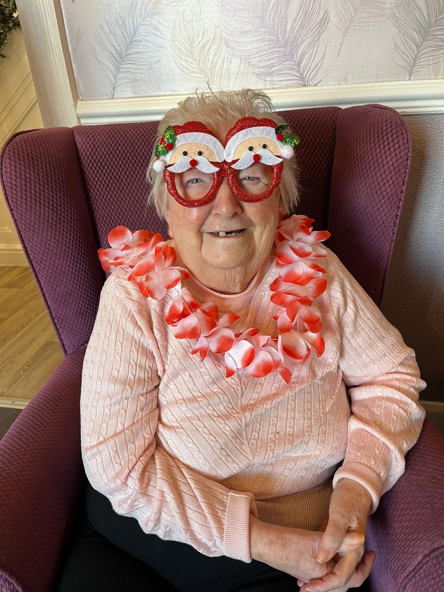 An elderly woman sitting in a purple armchair, wearing festive Christmas glasses shaped like Santa Claus and a pink lei, smiling.
