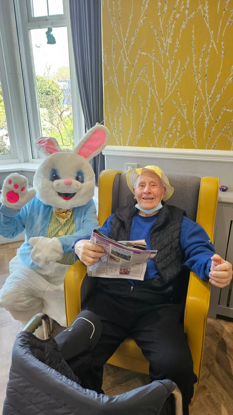 Person sitting in a yellow chair with a rabbit mascot standing beside them, both indoors near a window with curtains, a newspaper, and a walker nearby.