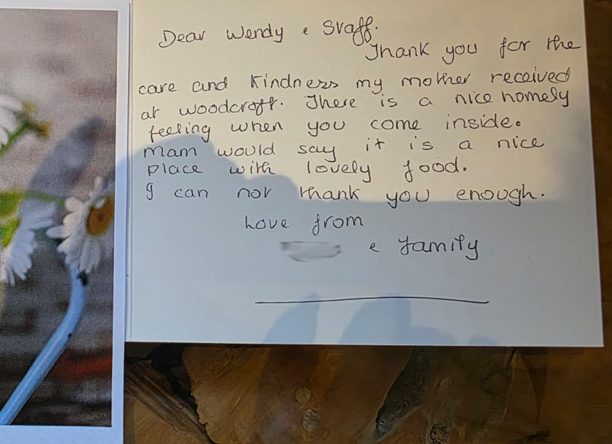 A handwritten thank you note on a yellow paper with an image of daisies and a partial view of a wooden surface.