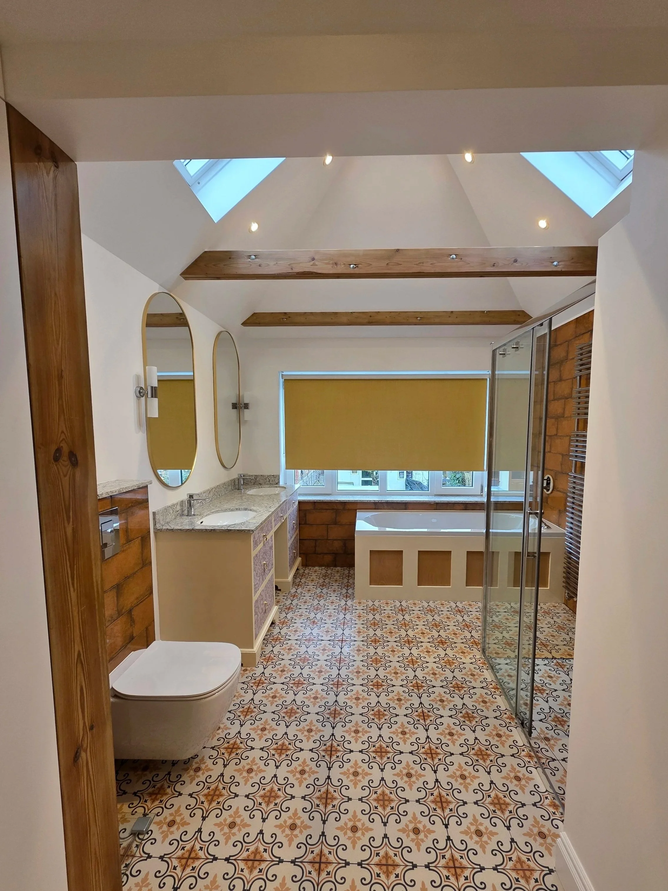 Bathroom with patterned tile floor, a double vanity with two mirrors, a toilet, and a walk-in shower with glass door. Skylights and window let in natural light.